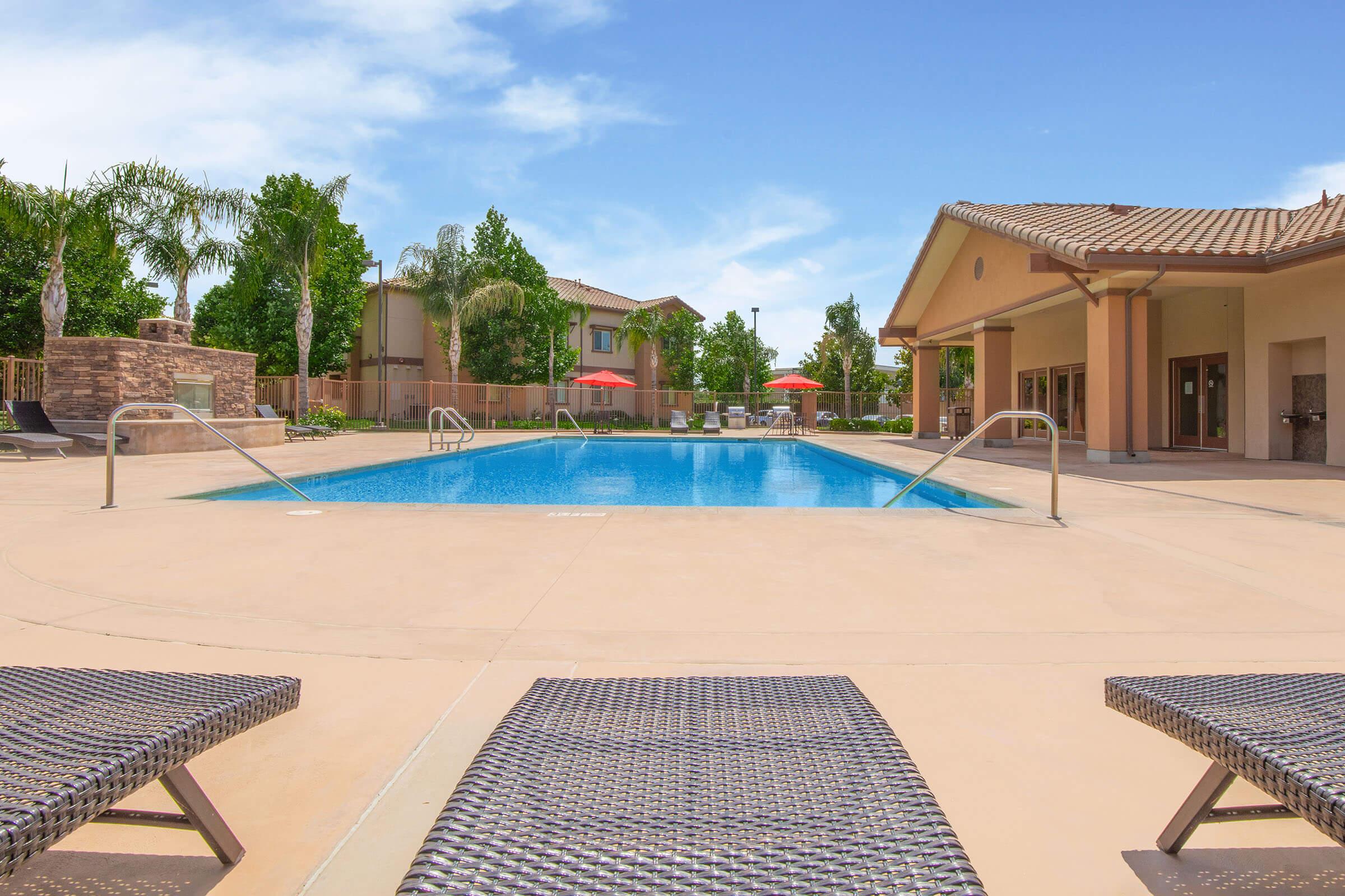 A tranquil outdoor swimming pool surrounded by lounge chairs, palm trees, and shaded lounge areas. A building with a covered patio and umbrellas is visible in the background, set against a clear blue sky. The scene conveys a relaxing atmosphere ideal for leisure and enjoyment.