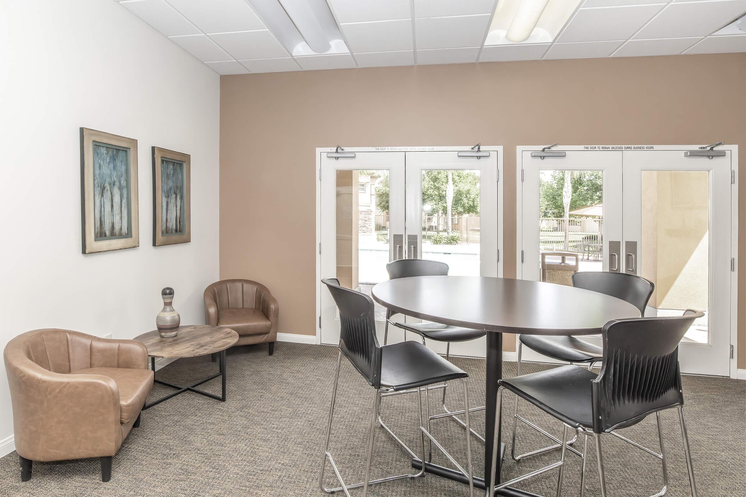 A modern, minimalist lounge area featuring two brown armchairs, a round table with high black chairs, and two sets of glass doors that open to an outdoor space. The walls are light beige, and there are two framed artworks on the wall. The flooring is carpeted, creating a cozy atmosphere.