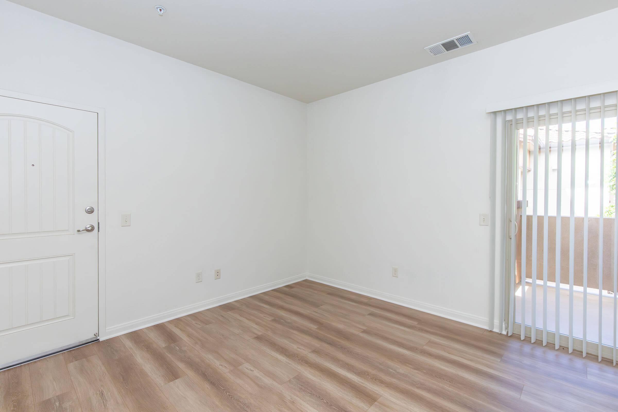 A vacant room with light-colored walls and wooden flooring. The room features a door on the left side and a sliding glass door with vertical blinds on the right, leading to a balcony or outdoor area. The space is bright and airy, suitable for various interior design options.