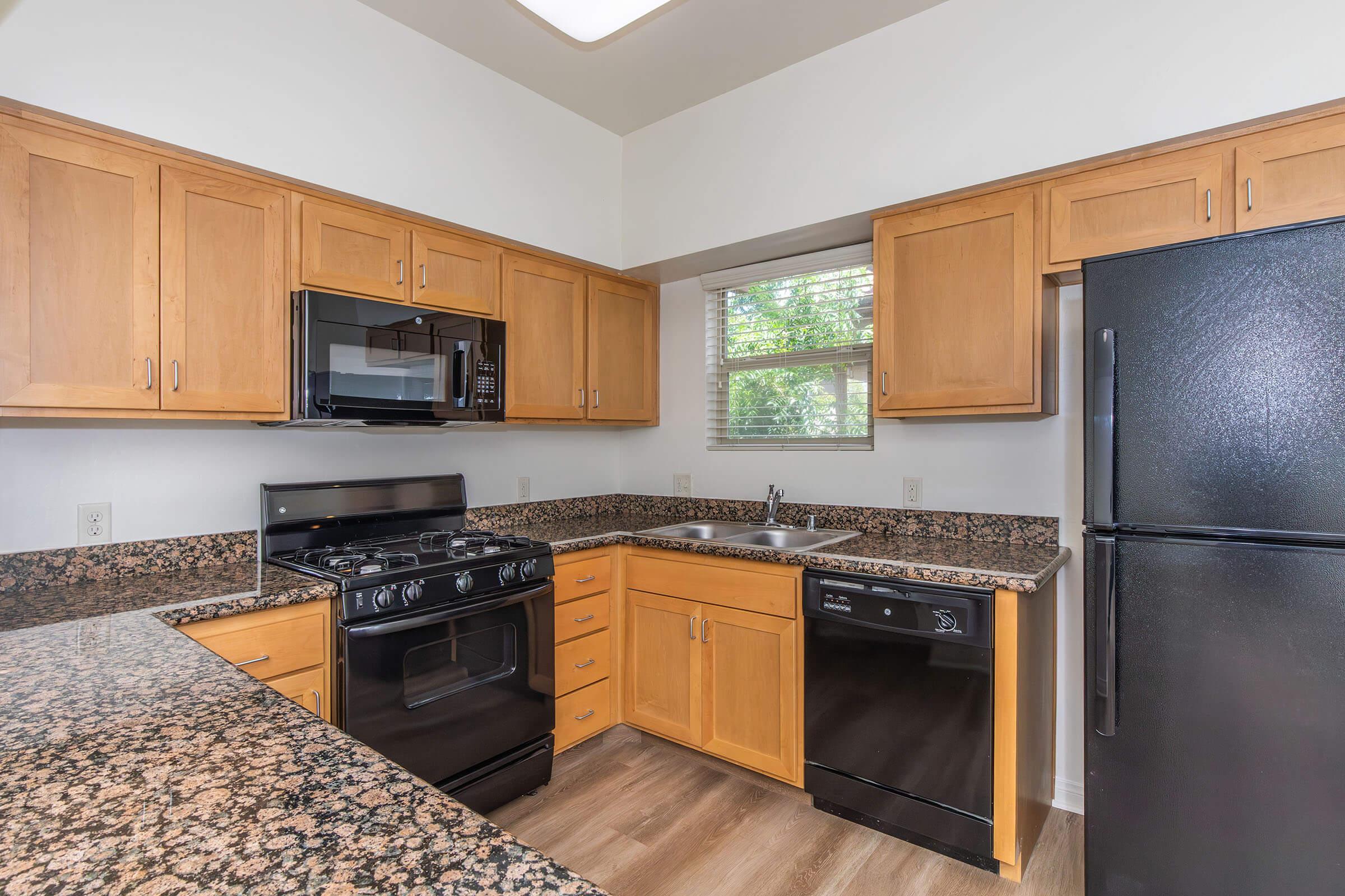 Modern kitchen featuring wooden cabinets, a granite countertop, stainless steel microwave, and gas stove. Includes a dishwasher and black refrigerator. A window provides natural light. The flooring is light-colored, adding to the contemporary look.