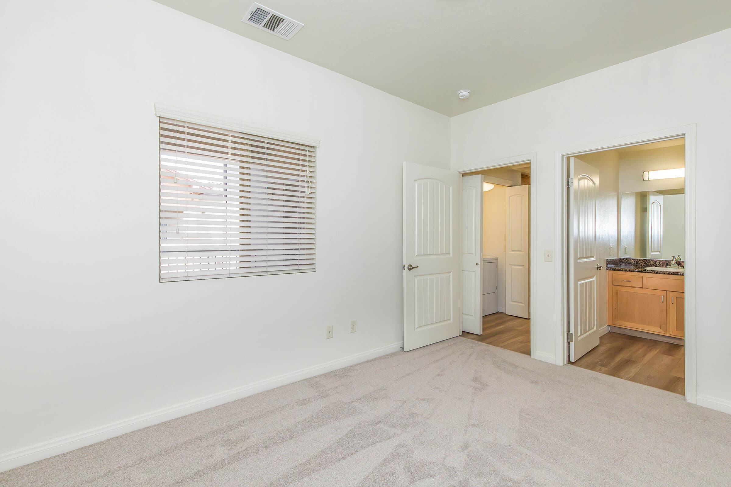 A lightly furnished room with white walls and beige carpet, featuring a window with blinds. Two doors lead to a bathroom and another area, with natural light coming in from the window. The space appears clean and welcoming, suitable for various living arrangements.