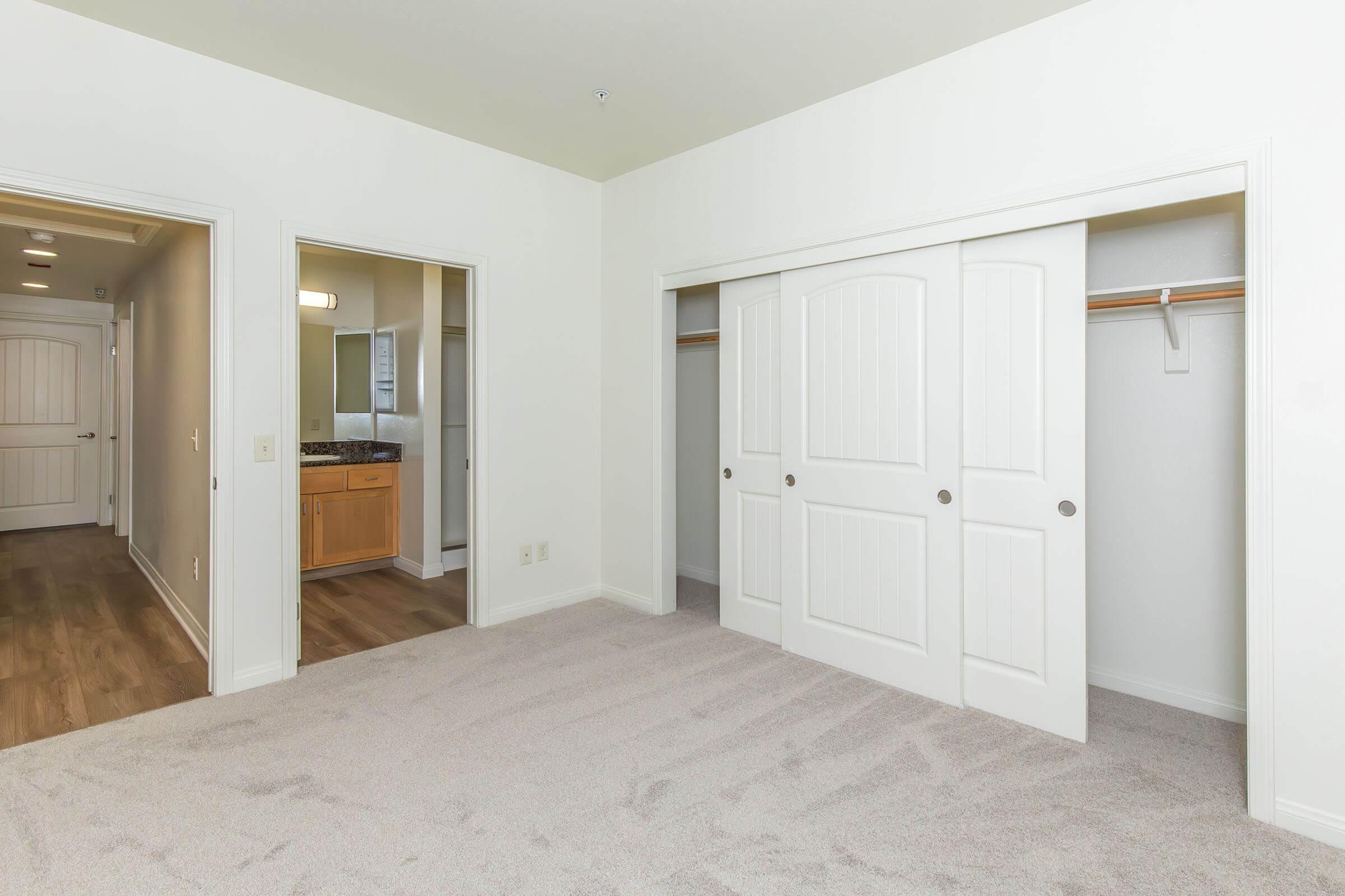 A spacious bedroom with soft, light-colored carpet and white walls. Two doorways lead to a bathroom and another room. A large closet with sliding double doors is visible, and the lighting is bright and inviting. The overall decor is modern and clean.