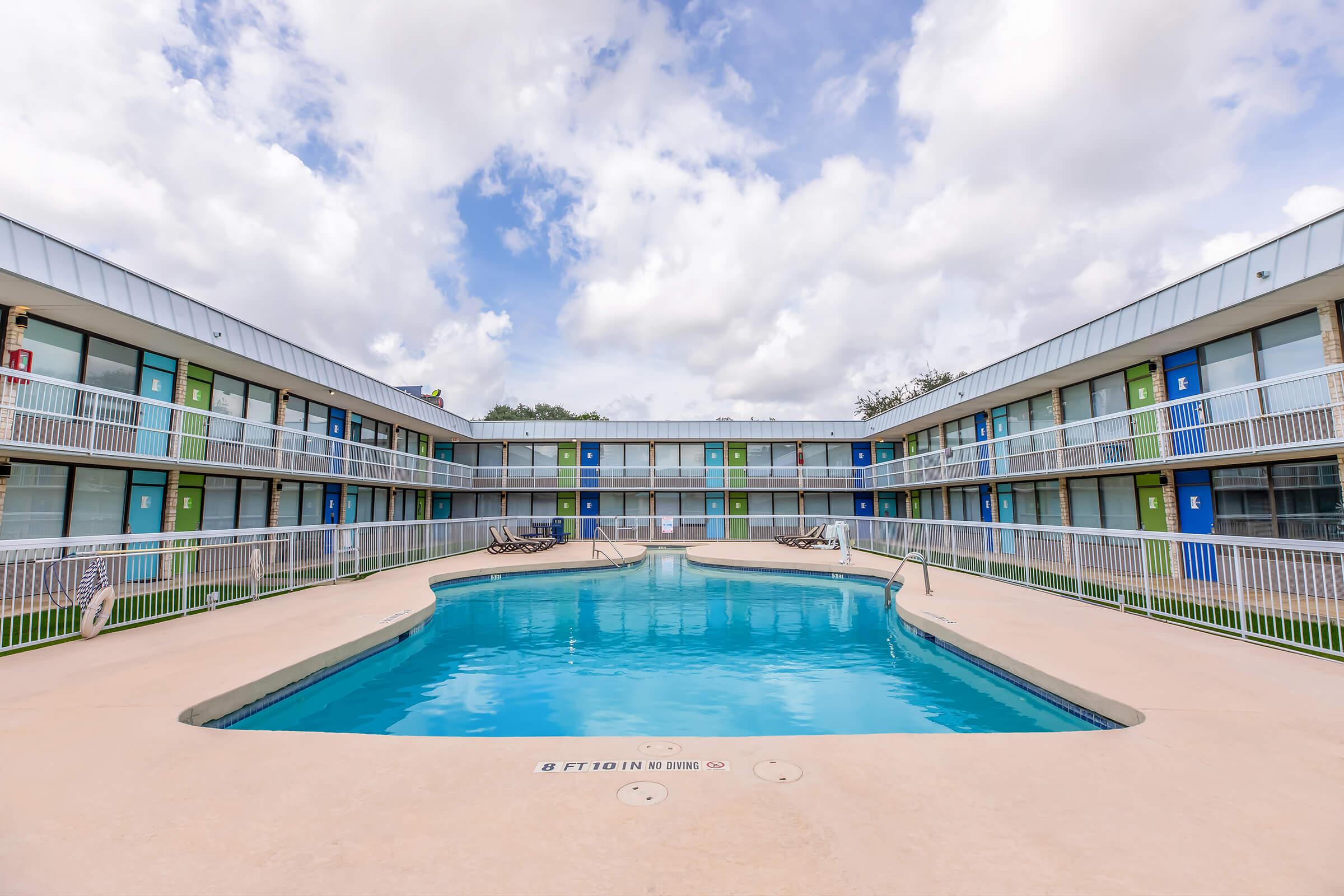 A rectangular swimming pool surrounded by a two-story motel with colorful doors. The pool area is paved, and there are lounge chairs on one side. The sky is partly cloudy, creating a bright atmosphere. The overall setting is inviting and designed for relaxation.