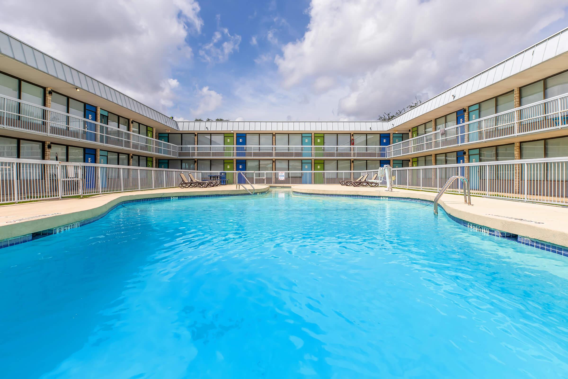 A clear blue swimming pool surrounded by a multi-story building with balconies. The structure features several colorful doors and windows. White railings border the pool area, and there are lounge chairs positioned nearby. The sky is partly cloudy, adding a bright and inviting atmosphere.