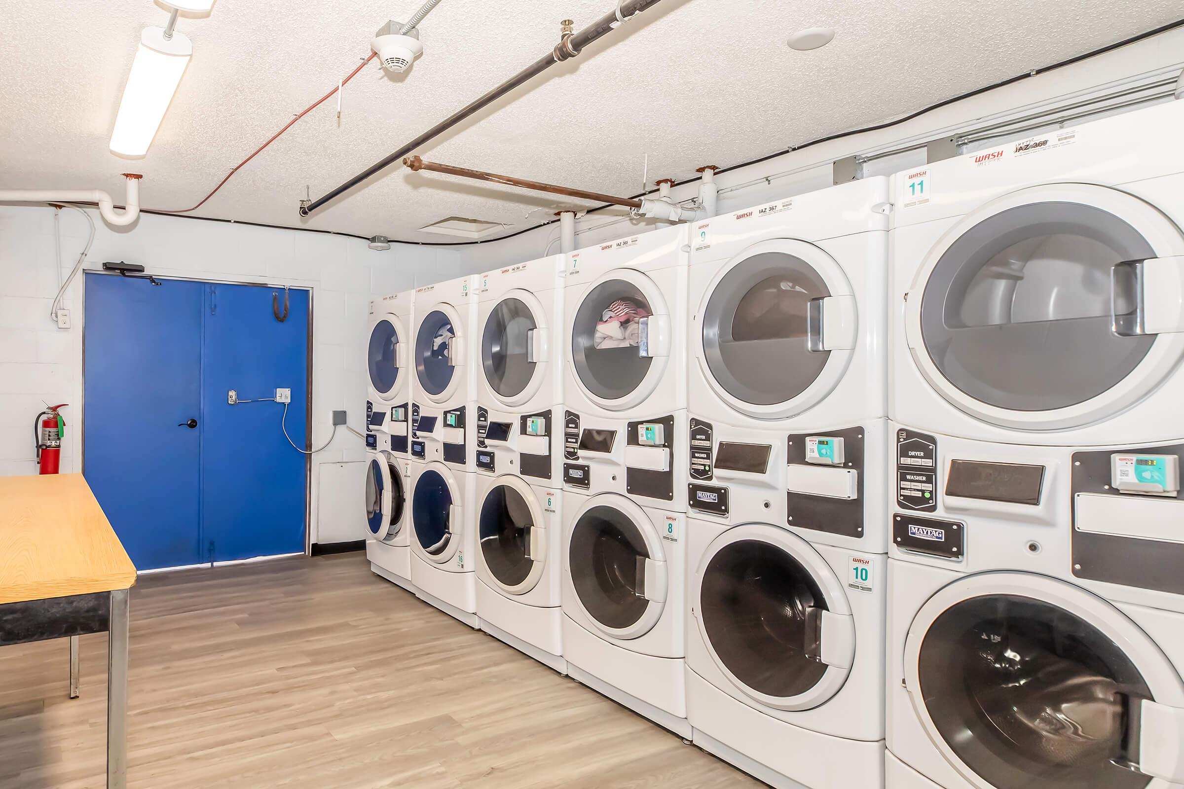 A laundry room featuring several white washing machines and dryers, lined against a wall. There is a blue door on one side and a table in front. The floor is wood-like and the ceiling has exposed pipes and fixtures.