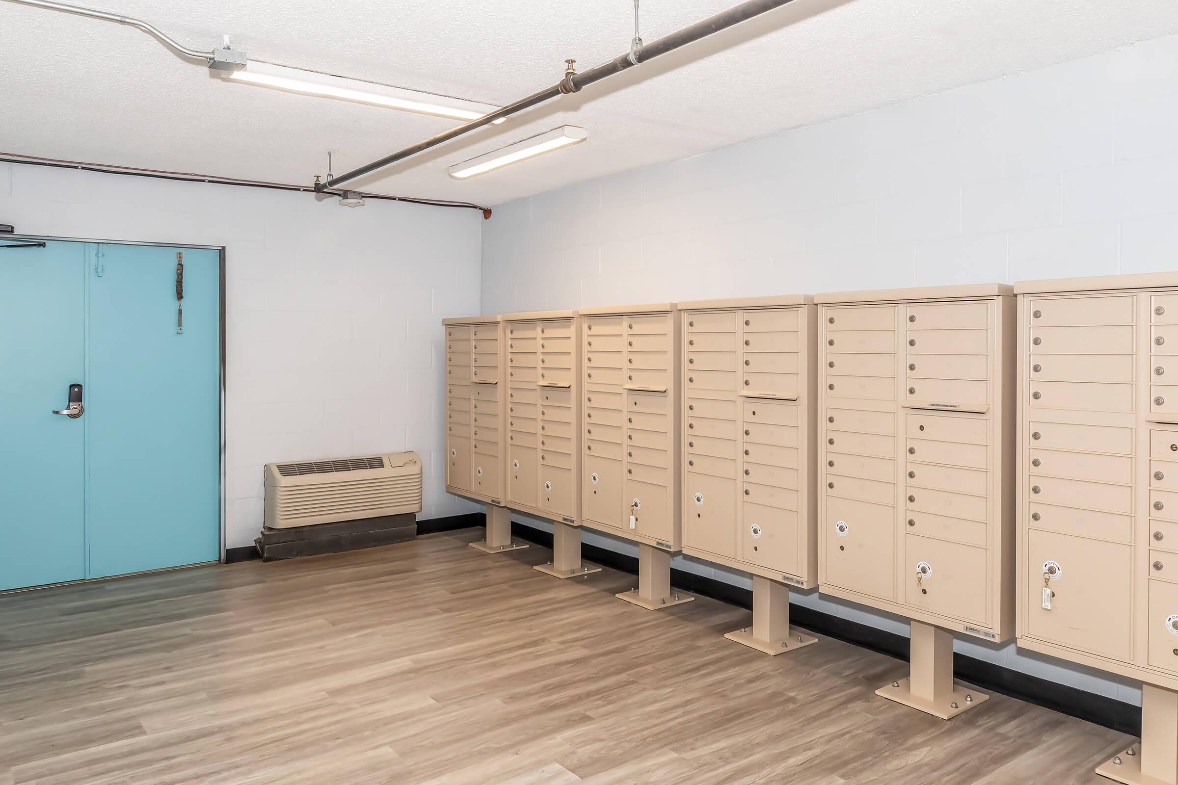 A clean, well-lit mailroom featuring a row of beige mailboxes with locks. A blue door is visible on one side, and an air conditioning unit is positioned against the wall. The floor is made of light wood-like material, contributing to a modern, tidy aesthetic.