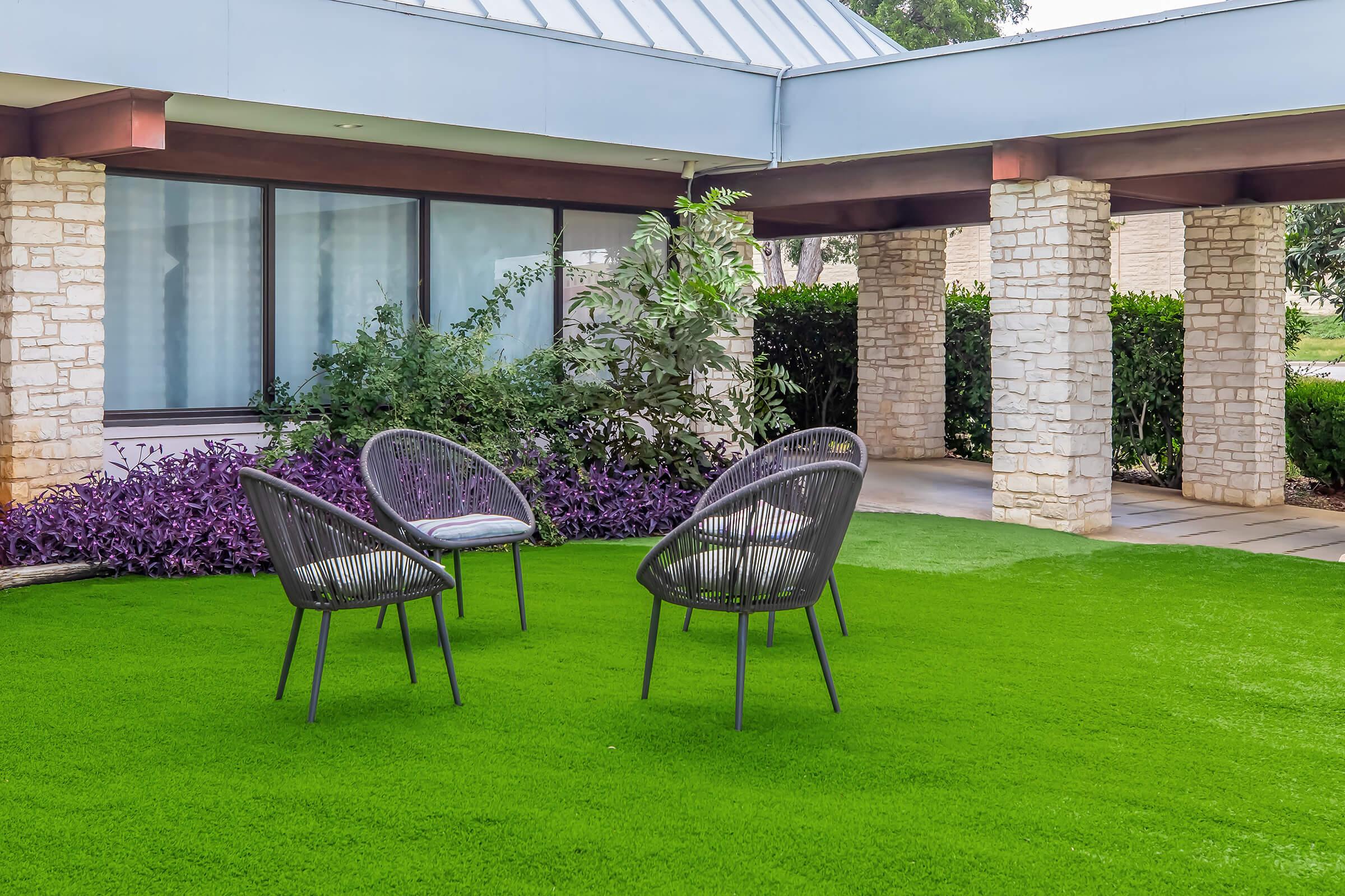 Three modern, woven chairs are arranged in a vibrant green artificial grass area, surrounded by decorative plants. The setting features a building with stone pillars and large windows, creating a serene outdoor space suitable for relaxation or socializing.