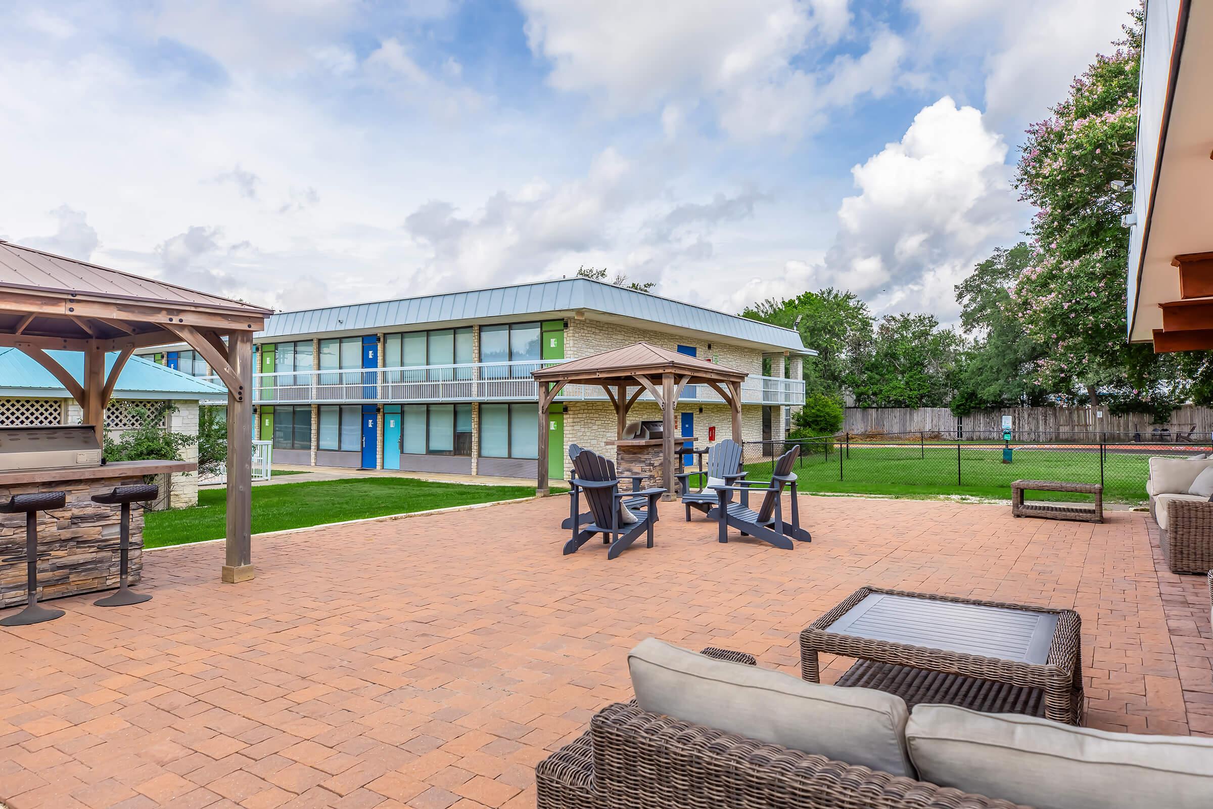 Outdoor area with a paved patio featuring lounge furniture and seating around a fire pit. In the background, a two-story building with blue and green accents is visible, surrounded by grass and trees, under a cloudy sky.