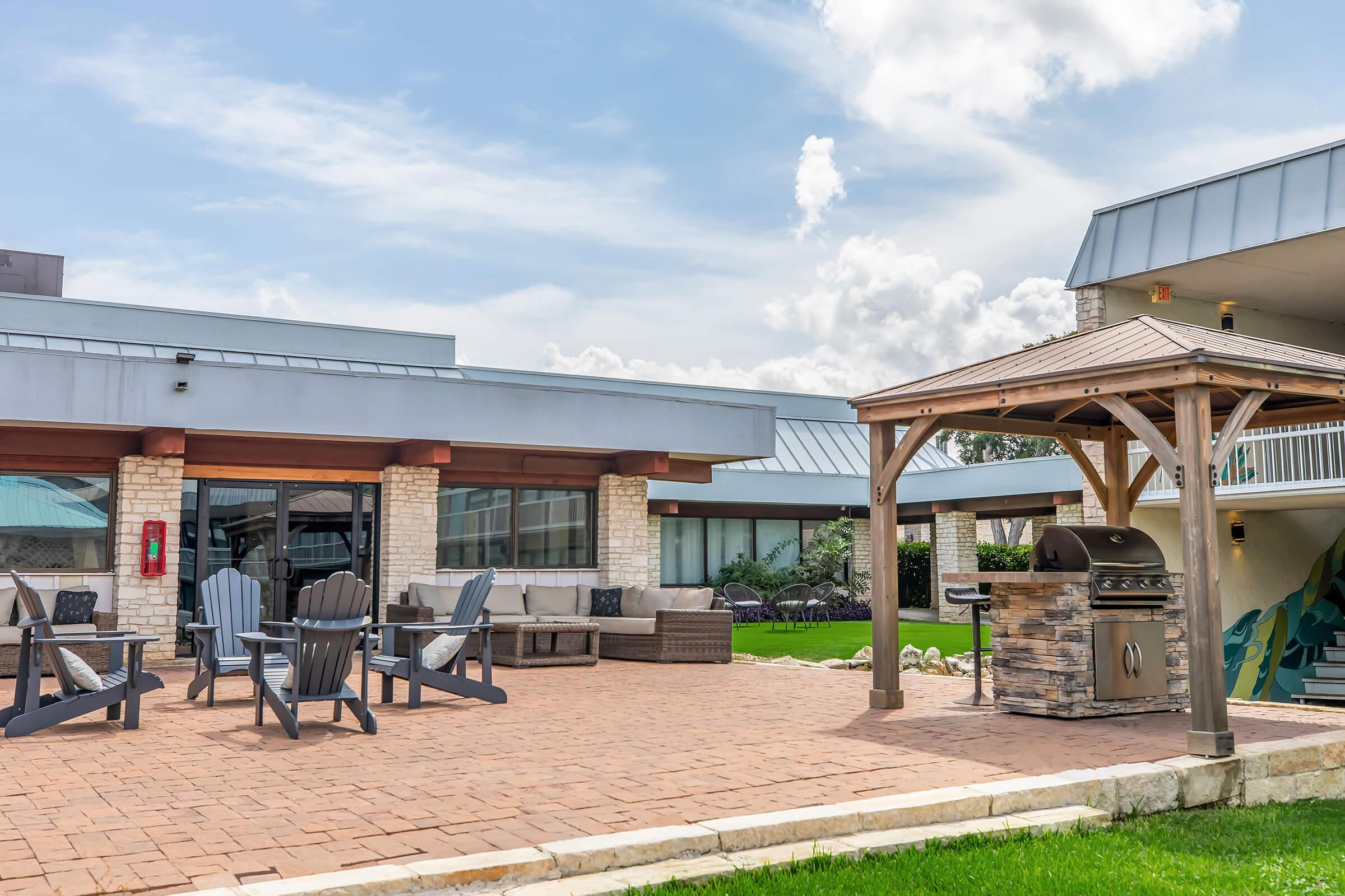 Outdoor patio area featuring a stone barbecue grill under a gazebo, surrounded by wooden lounge chairs and a comfortable seating arrangement. The setting is bright with blue skies and fluffy clouds, creating a welcoming atmosphere for relaxation and outdoor cooking.