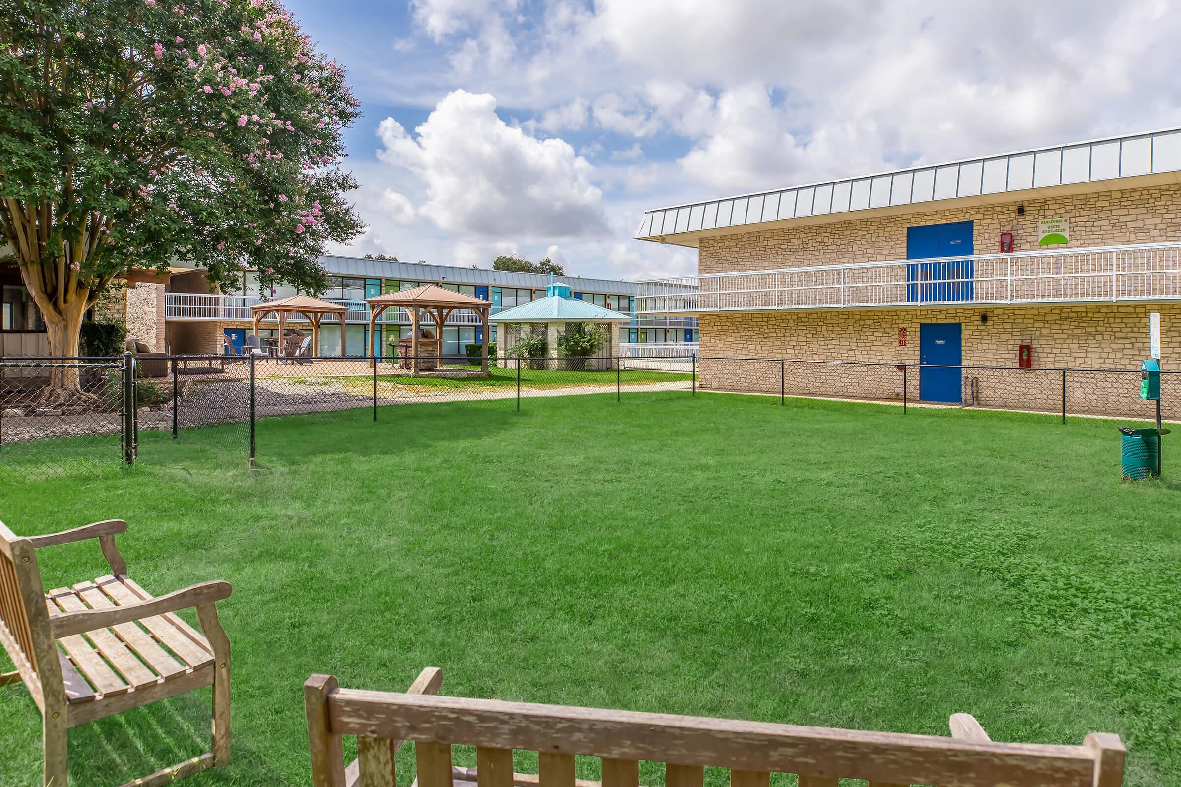 A grassy courtyard surrounded by brick and glass buildings, featuring wooden benches and shaded gazebo areas. Bright blue skies with scattered clouds above enhance the inviting atmosphere. The area is fenced, with pathways and green landscaping, creating a pleasant outdoor space for relaxation.