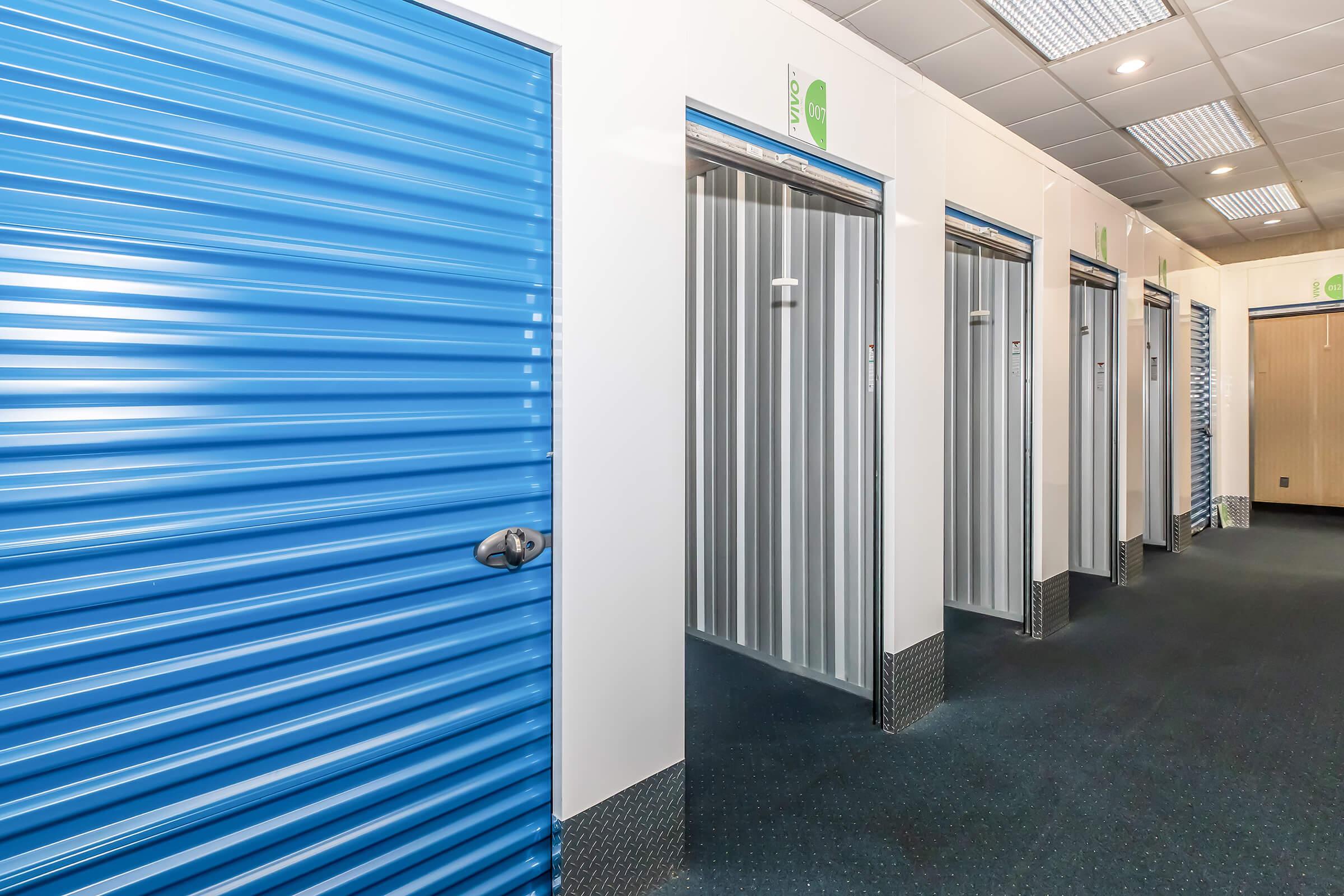 Interior view of a storage facility featuring several storage units with blue and silver metal doors. The hallway is well-lit and has a clean, modern appearance, with a dark carpeted floor and white walls. The storage units are neatly aligned, providing a clear and organized look.
