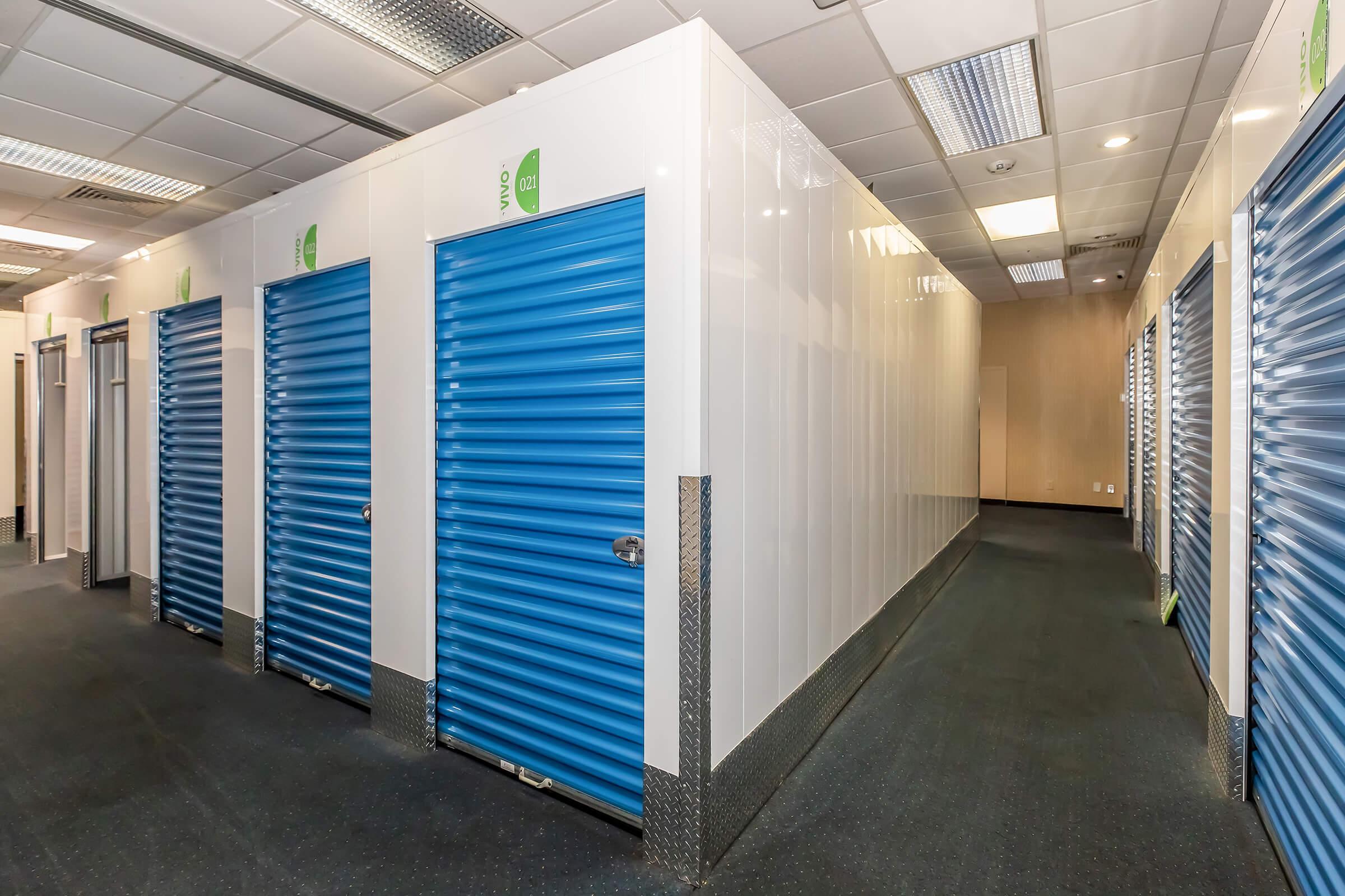 Interior view of a storage facility featuring several storage units with blue roll-up doors. The corridor is well-lit, with white walls and a dark floor, creating a clean and organized environment for storing items.