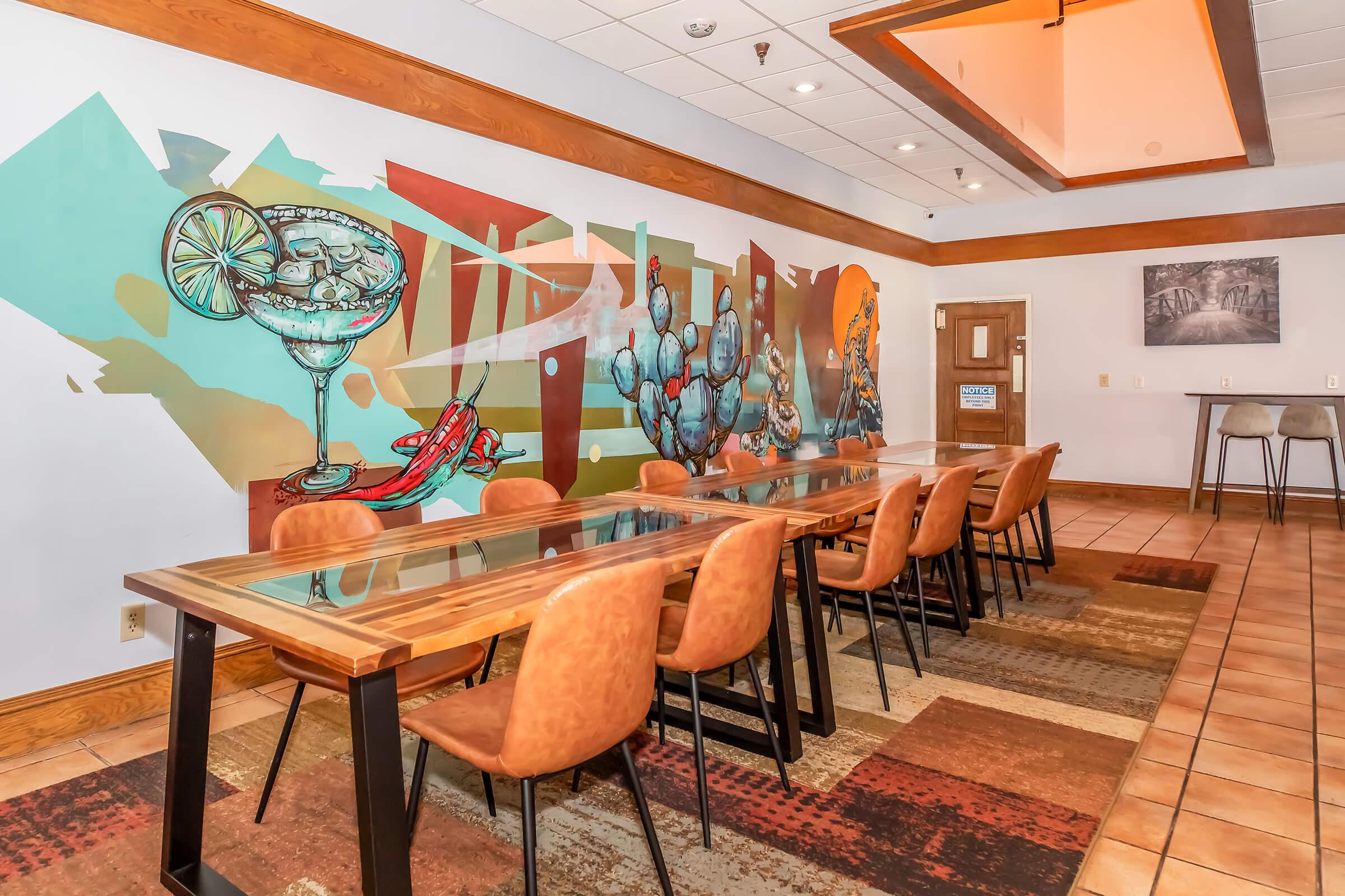 A modern dining area featuring a long wooden table surrounded by brown chairs. The wall is decorated with a colorful mural depicting cocktails, cacti, and artistic elements. The floor is covered with a patterned rug, and there is a smaller table in the background with high chairs. Brightly lit and inviting atmosphere.