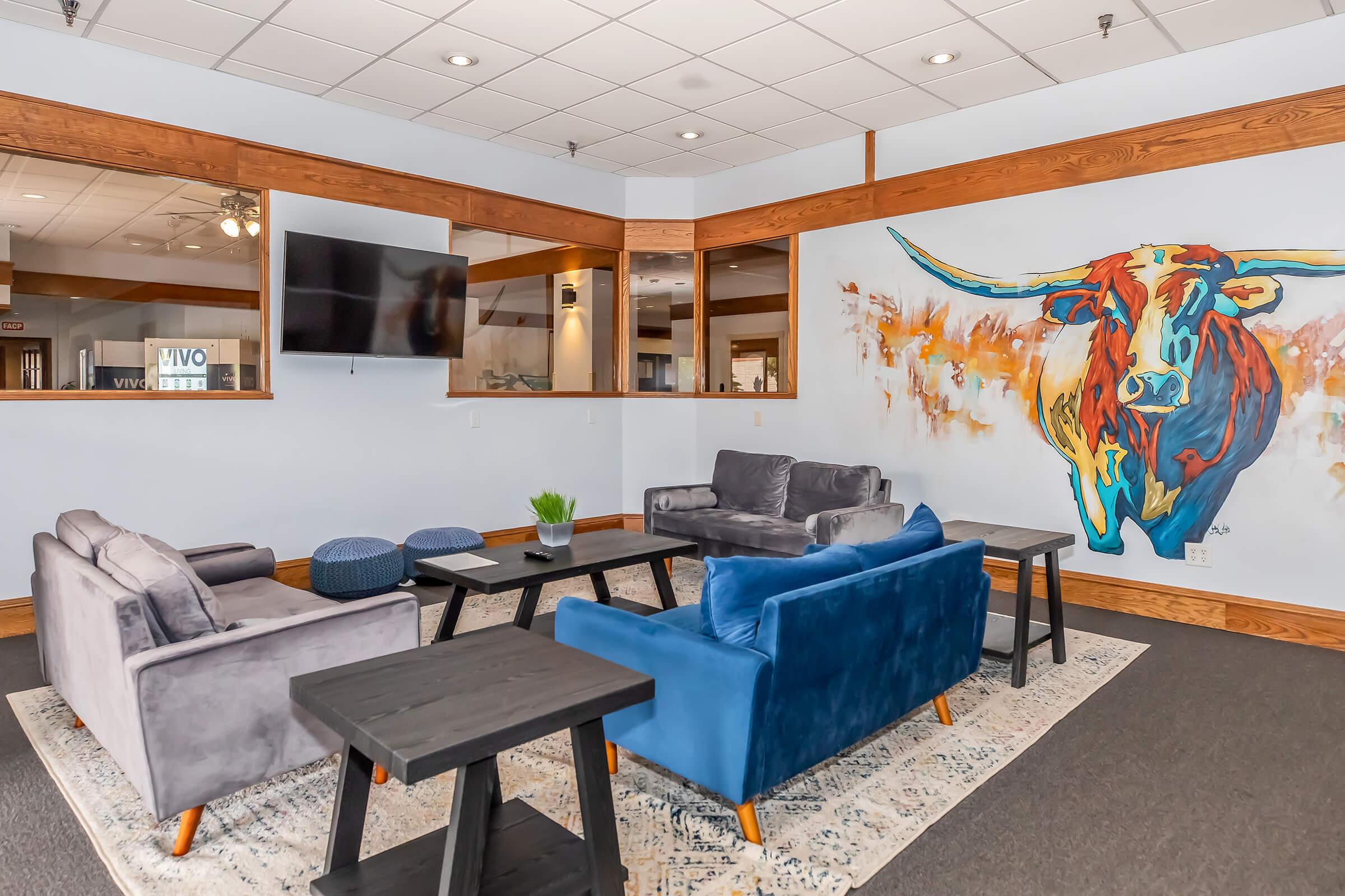 A cozy lounge area featuring a large colorful painting of a longhorn steer on the wall. Furnished with plush gray and blue sofas, a dark wooden coffee table, and small side tables, the space is designed for relaxation. A flat-screen TV is mounted on the wall in the background.