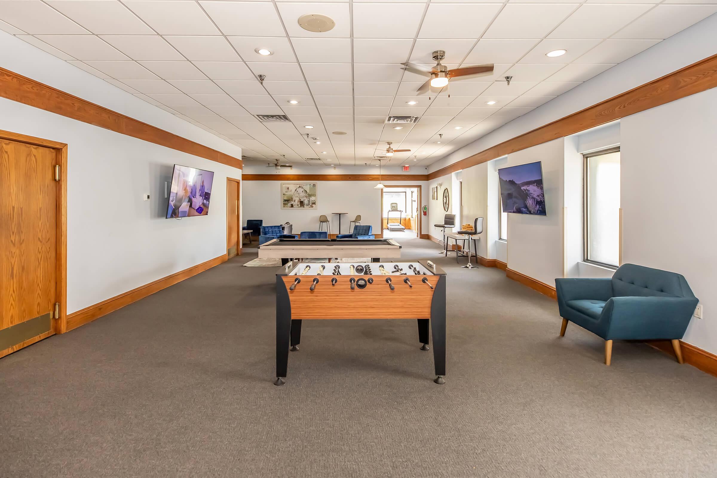 A spacious, well-lit room with a foosball table in the center. There are two large flat-screen TVs mounted on the walls, comfortable seating areas, and a variety of tables. The flooring is carpeted, and the walls are adorned with light colors, creating a welcoming atmosphere.