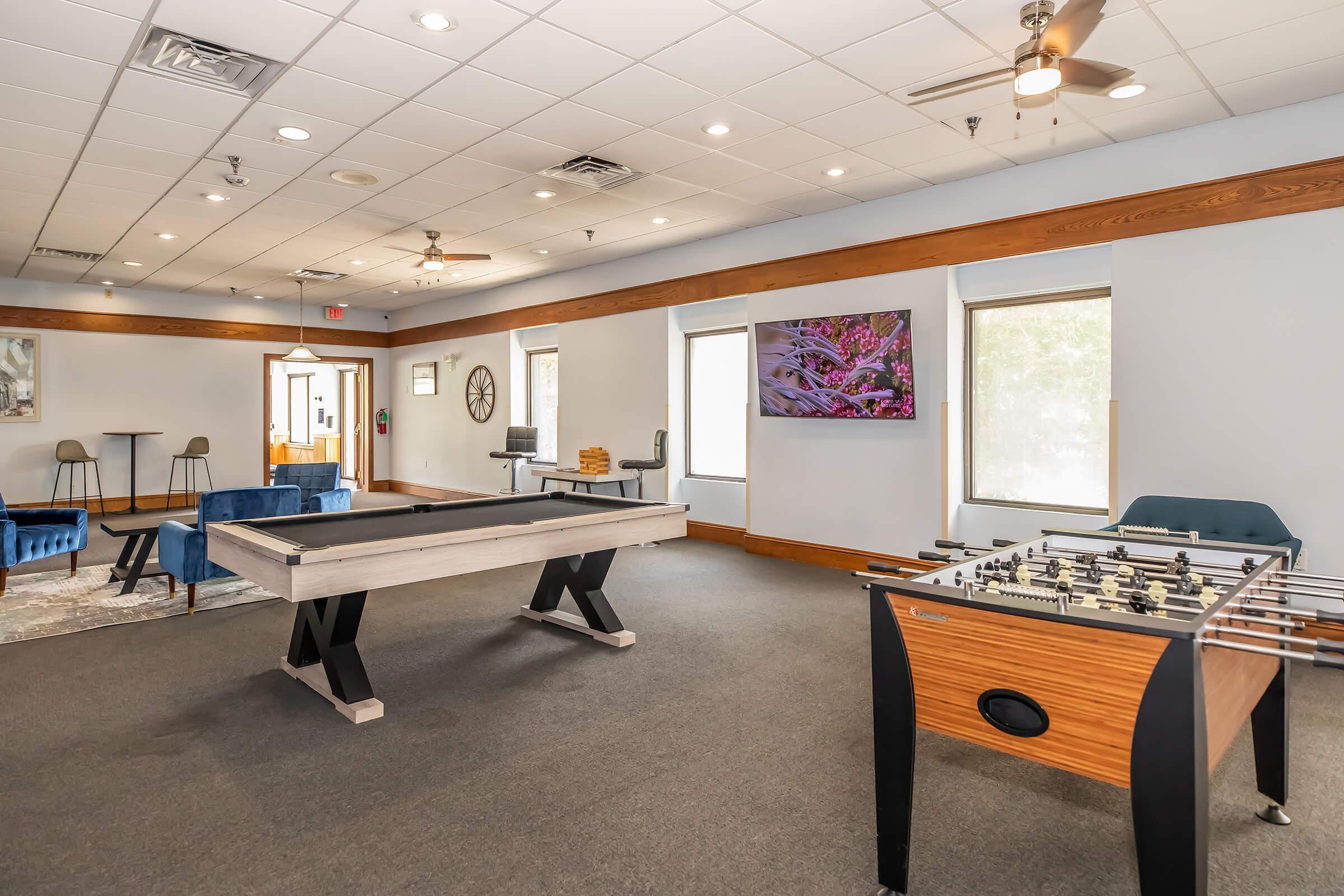 A spacious recreational room featuring a pool table and a foosball table. Comfortable seating areas with blue chairs and a small table are visible. A large wall-mounted TV displays vibrant artwork. The room has a clean, modern design with ample natural light from windows.