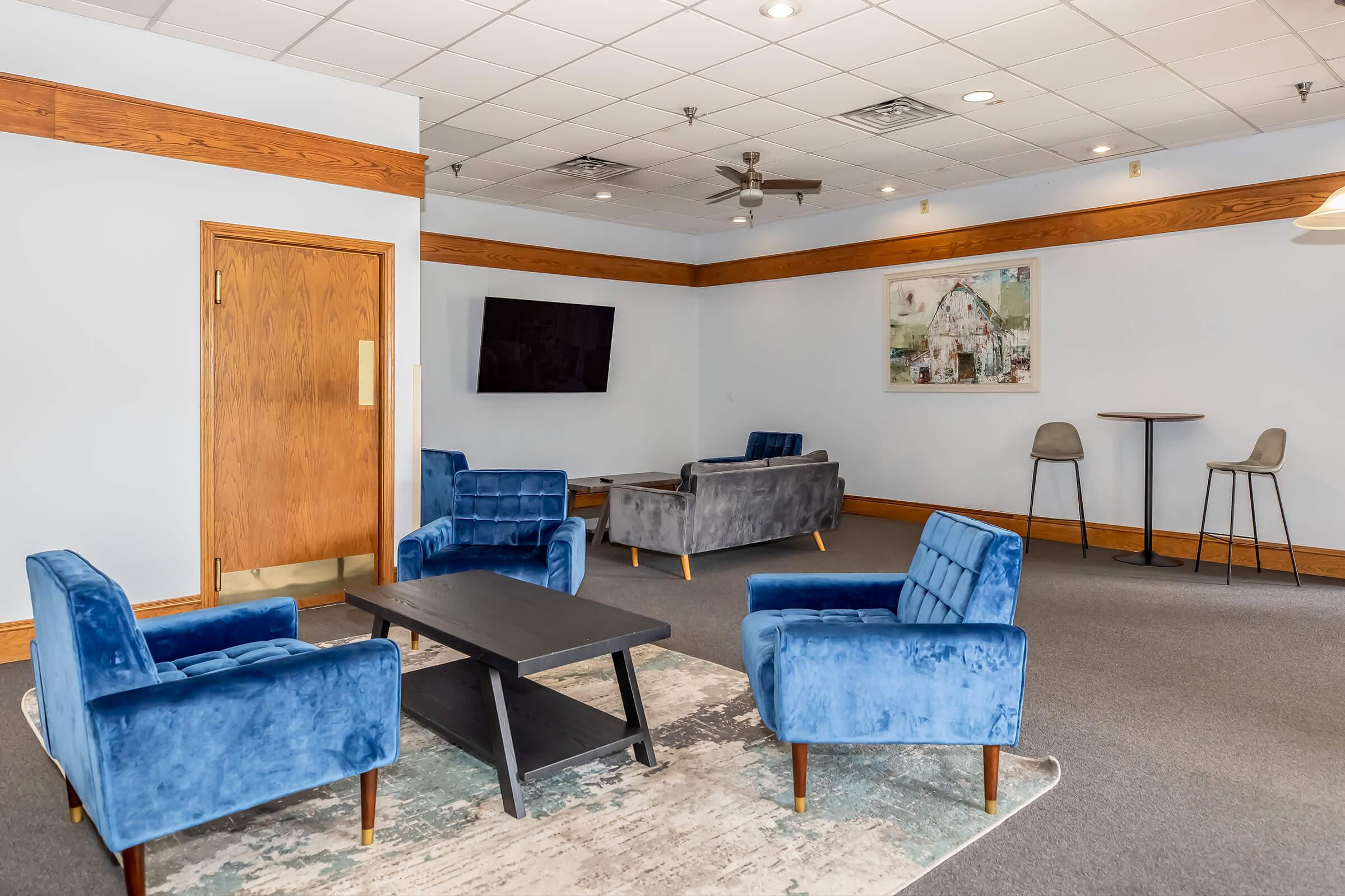 A modern lounge area featuring blue upholstered chairs, a dark coffee table, and a flat-screen TV mounted on the wall. The walls are painted light blue, and there’s a piece of art depicting a rustic building. A small high table with stools is visible in the background. The space has a relaxed atmosphere.