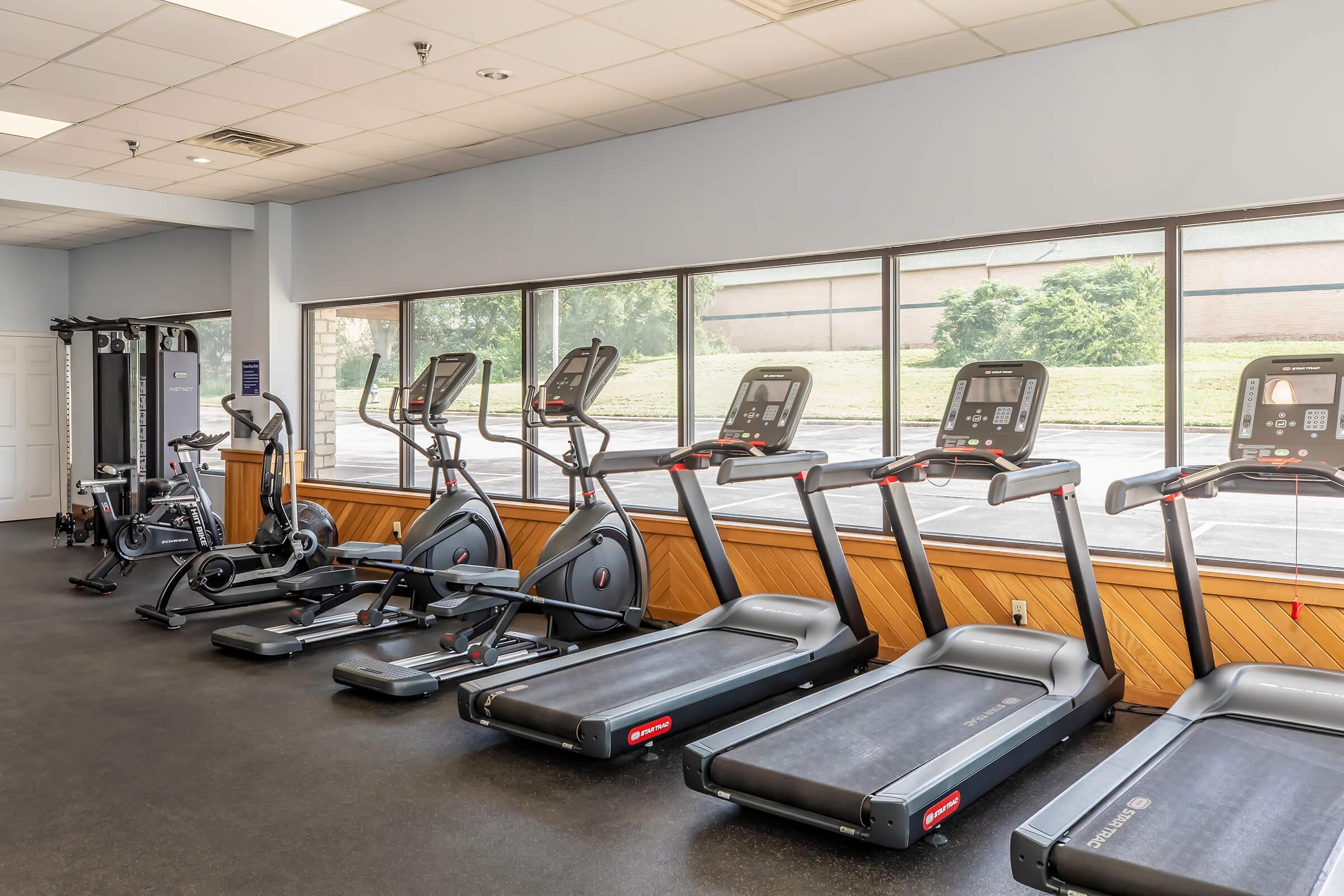 A bright and spacious gym with various exercise equipment. The room features multiple treadmills, an elliptical machine, and a stationary bike, all arranged neatly against large windows that provide a view of the outdoors. The flooring is dark, enhancing the modern gym atmosphere.