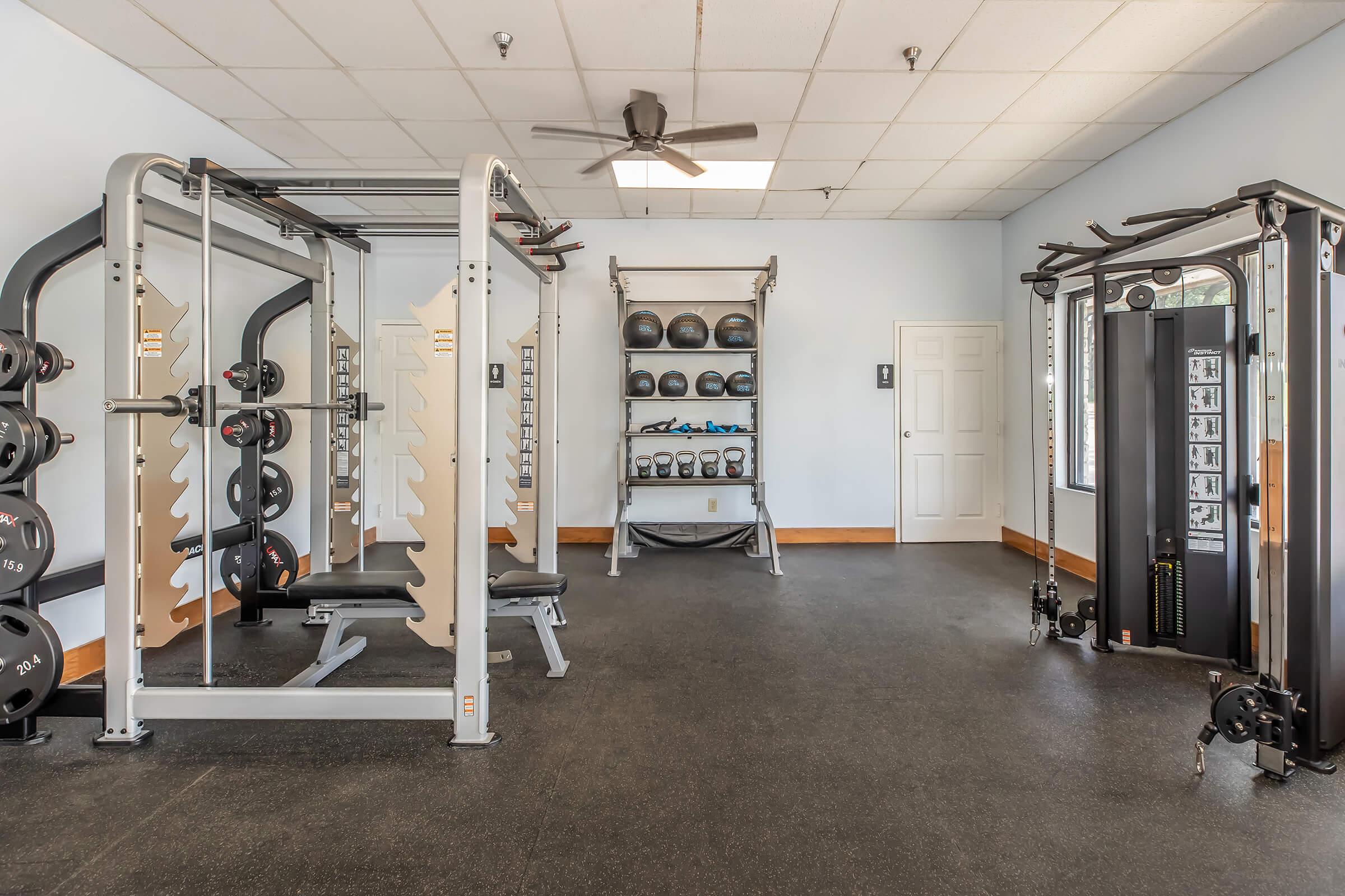 A spacious gym interior featuring weightlifting equipment, including a multi-station weight machine, free weights on a rack, and a bench press area. The room has a ceiling fan, bright lighting, and a light-colored wall. There is also a door leading outside.