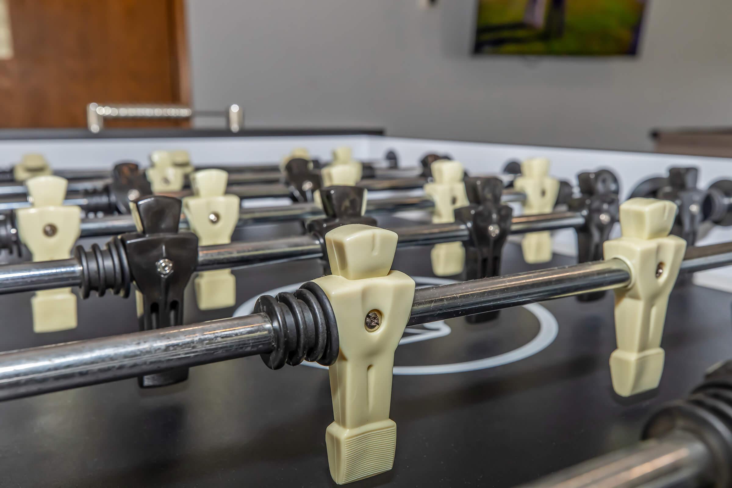 A close-up view of a foosball table featuring rows of players in black and beige colors mounted on metal rods. The background includes a blurred image of a TV screen and a wooden wall. The scene captures the excitement of a foosball game with a focus on the player figures.
