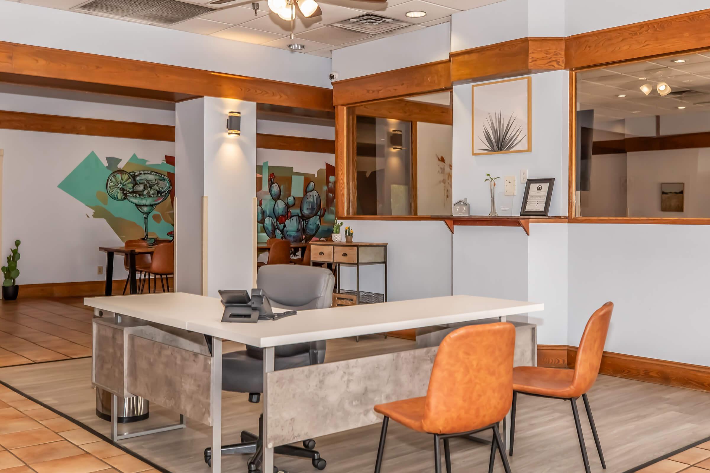 A modern office reception area featuring a large, minimalist desk with a telephone, a brown chair, and a decorative wall with plants and artwork. Light-colored walls and wooden accents create a warm atmosphere. There are additional chairs and a reflection in the mirror, enhancing the space.