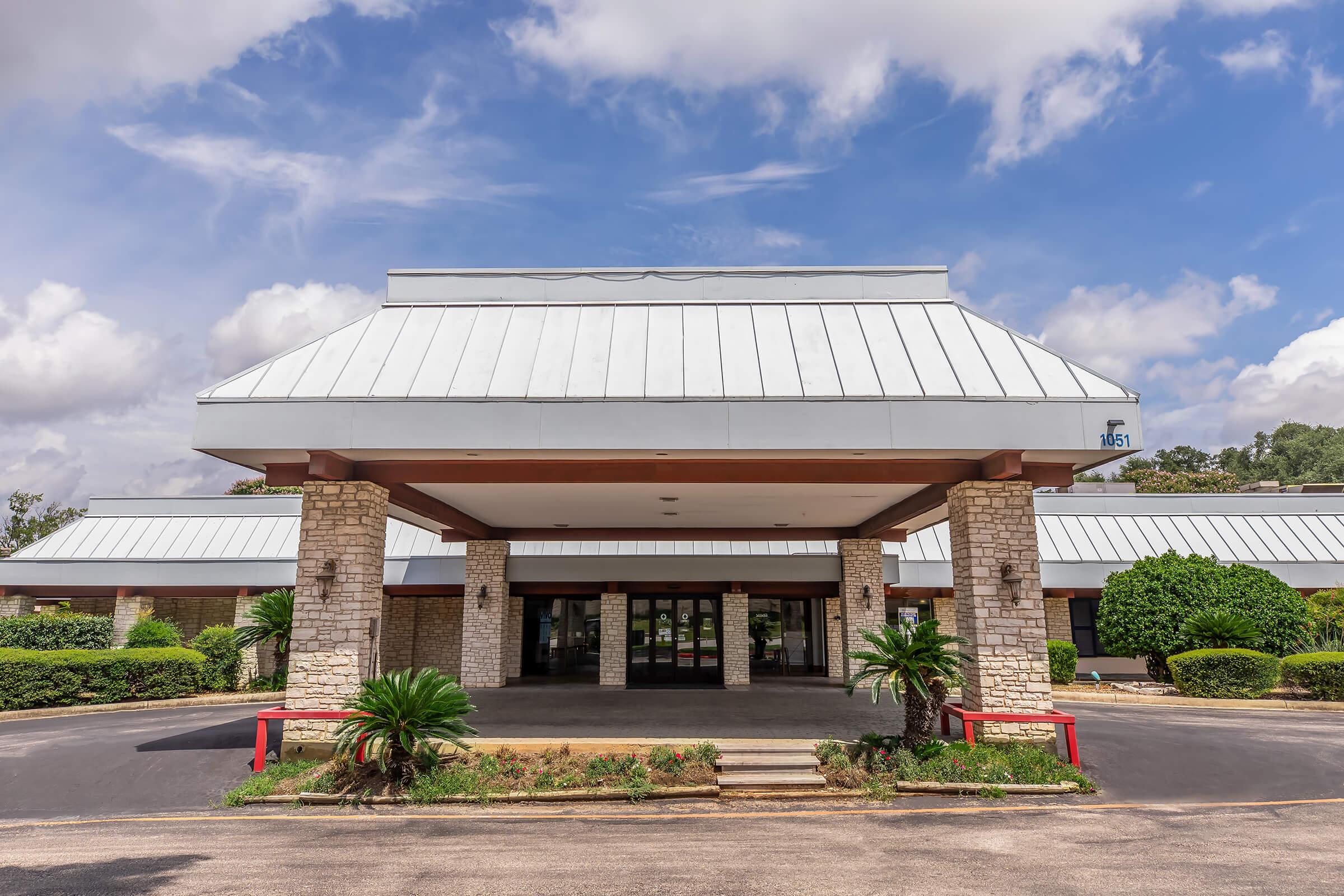 A single-story building with a modern, sloped roof and stone pillars at the entrance. The façade features large glass doors, flanked by lush greenery and palm plants. The sky above is partly cloudy, creating a bright, inviting atmosphere. The parking area in front is paved and well-maintained.