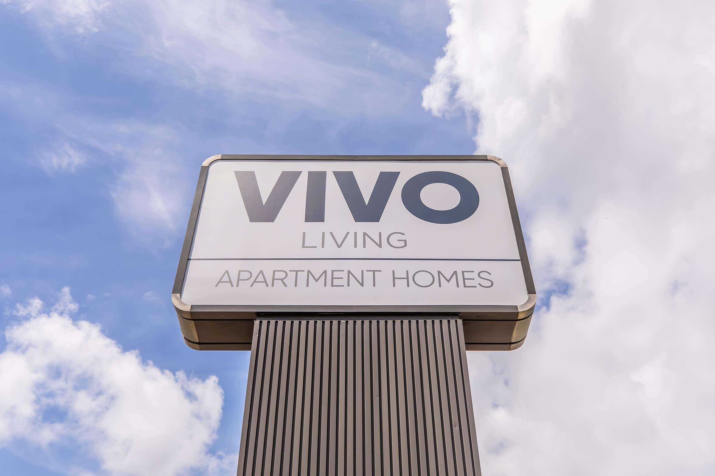 Sign for "VIVO Living Apartment Homes" with a clear blue sky and fluffy clouds in the background. The sign is prominently displayed, featuring the name "VIVO" in large letters.