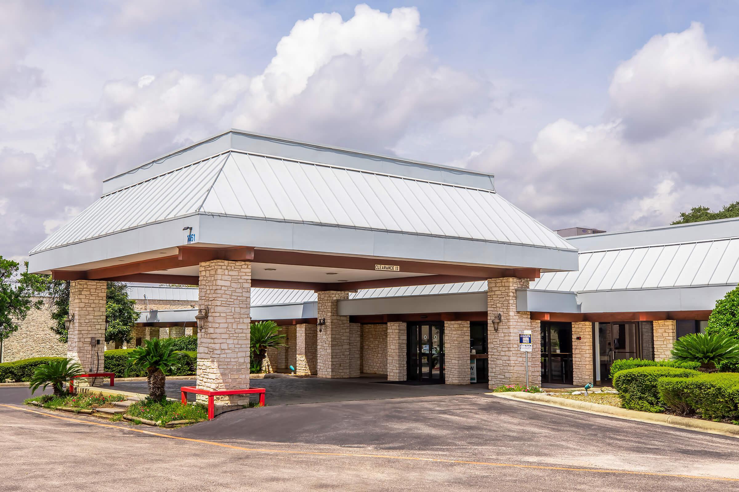 A modern building entrance featuring a low, flat-roof design with a metal roof. The facade is made of light-colored stone, complemented by wooden beams. Surrounding the entrance are landscaped areas with palm trees and shrubs, under a partly cloudy sky. There are parking spaces visible in front of the entrance.