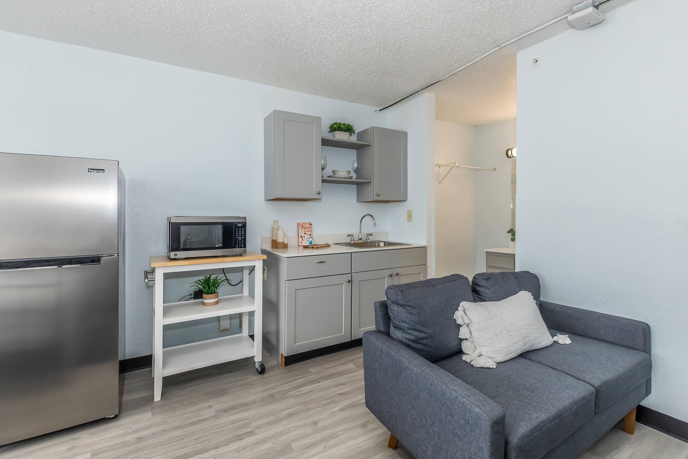 A modern kitchenette featuring a stainless steel refrigerator, microwave, and a small sink. There is a white cart with kitchen essentials and a cozy gray sofa with a decorative throw pillow. The walls are painted light blue, and there is a closet area visible in the background. The flooring is light wood.