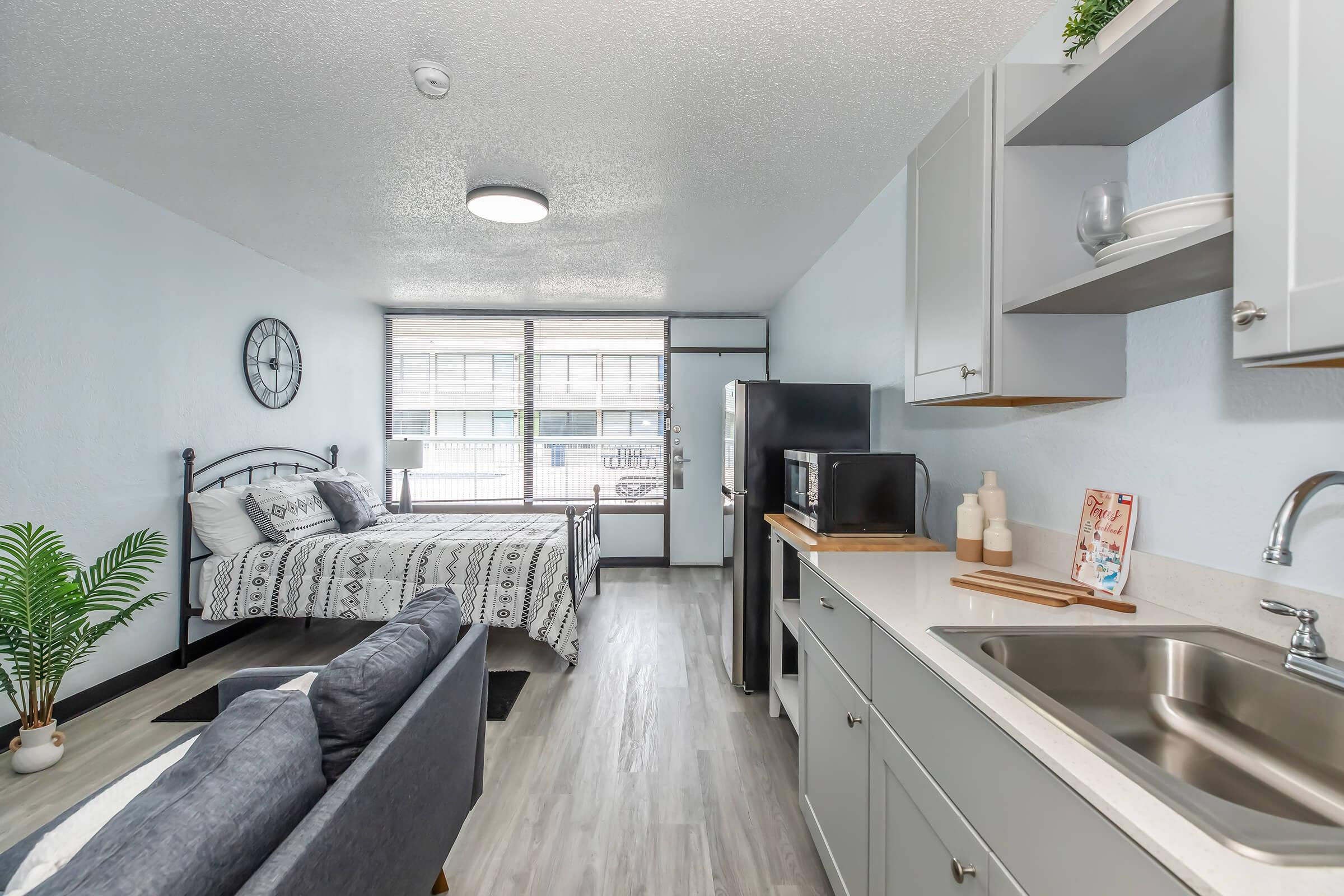A modern studio apartment featuring a double bed with patterned bedding, a small kitchenette with a sink and microwave, and a cozy couch. Large windows allow natural light to fill the space, and there is a decorative plant near the bed. The walls are painted light blue, creating a fresh atmosphere.