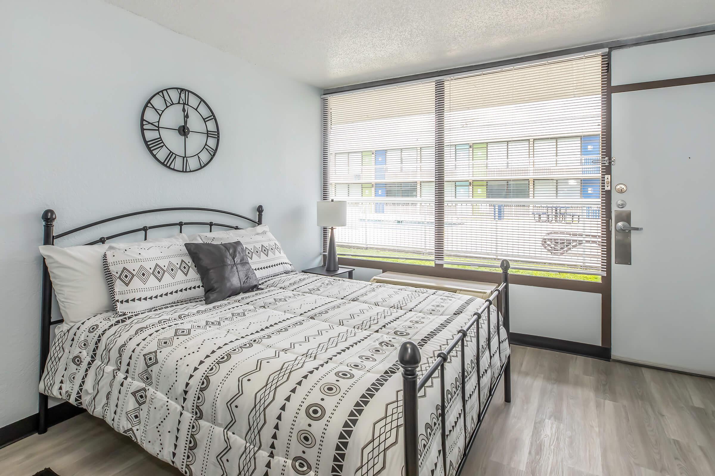 A cozy bedroom featuring a black metal bed frame with a patterned comforter and decorative pillows. A lamp sits on a small side table next to the bed. Large windows with blinds overlook a grassy area, and a wall clock adds a modern touch to the decor.
