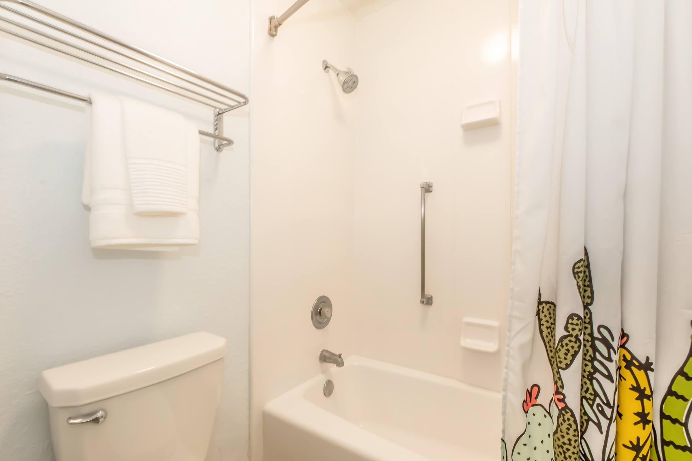 A clean and simple bathroom featuring a shower with a curtain adorned with colorful cacti, a white bathtub, a chrome grab bar, a sink faucet, and a toilet. Fresh towels are hanging on a rack nearby, against a light-colored wall, creating a bright and inviting atmosphere.