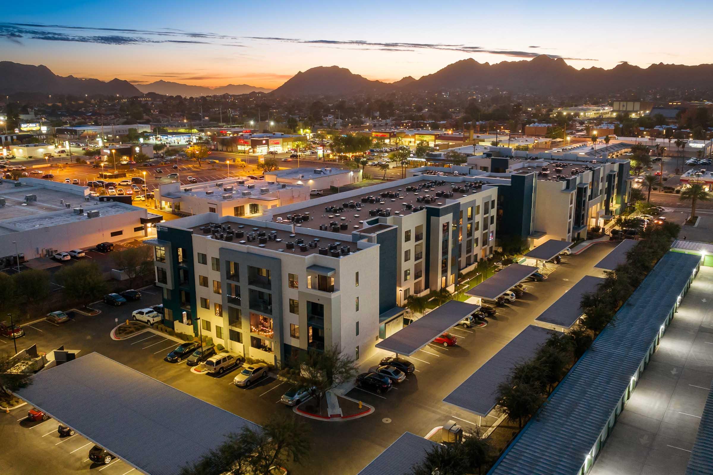 Aerial view of a modern apartment complex at sunset, surrounded by a commercial area. 