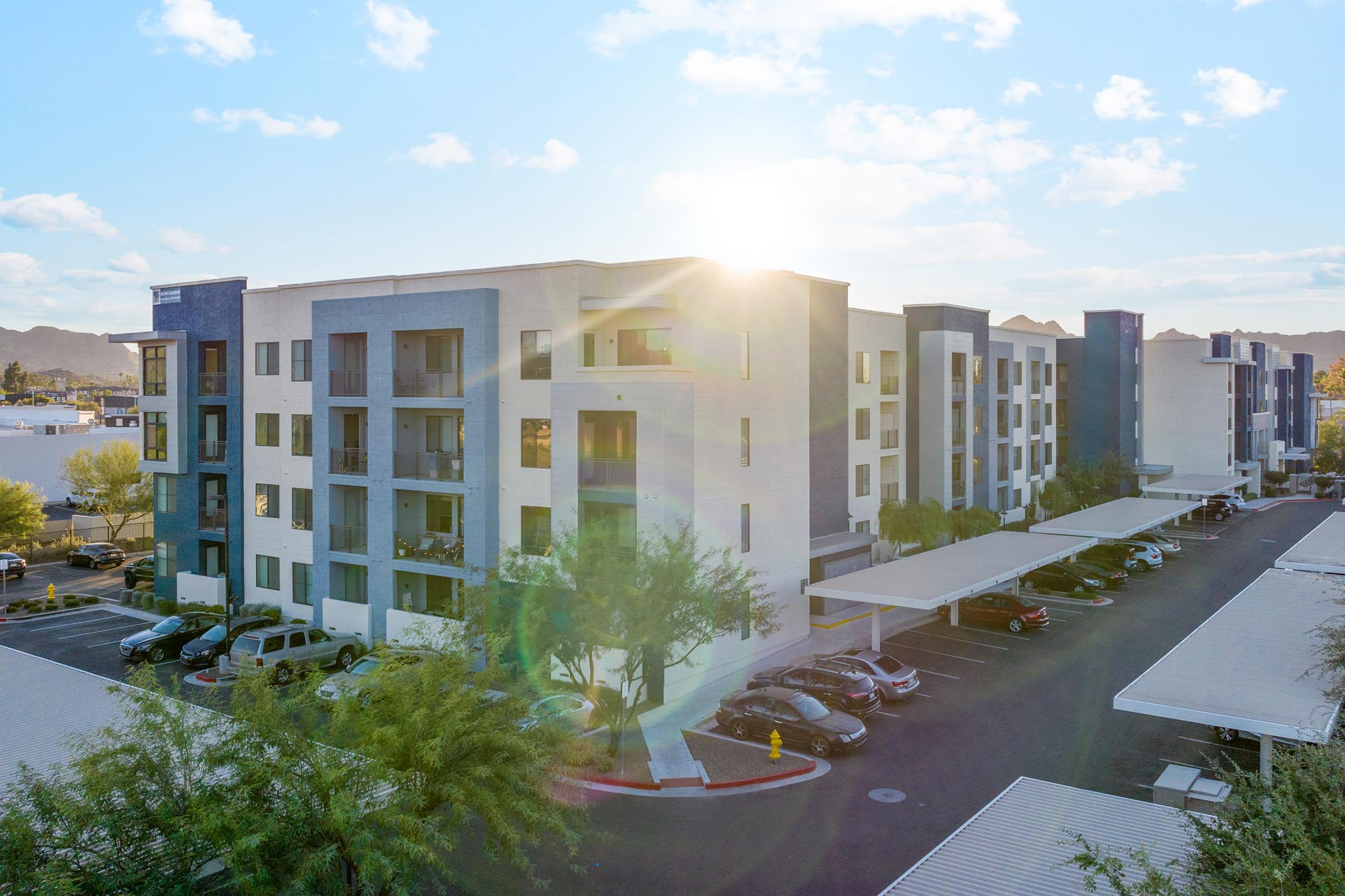 Modern apartment building with a light-colored facade and balconies, situated in an outdoor parking area with shaded spots. 