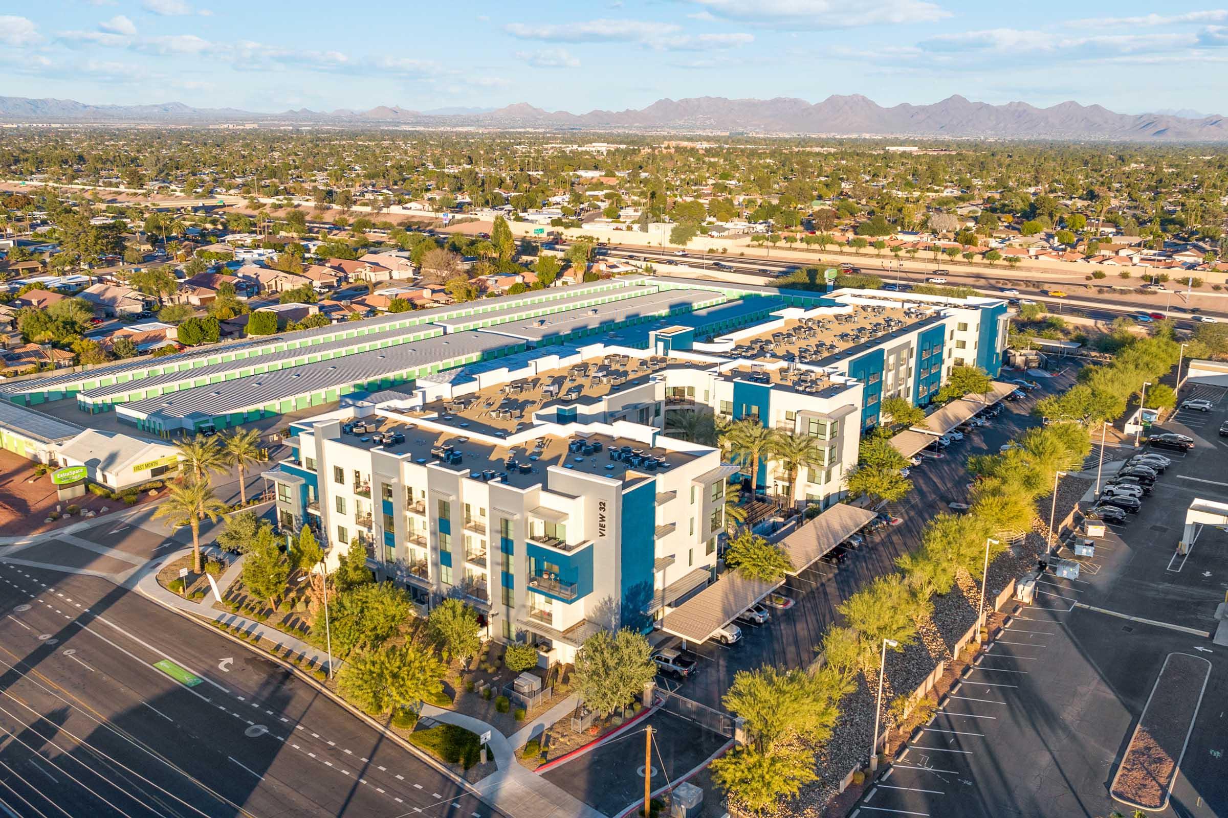 Aerial view of a modern multi-story apartment building featuring blue and white accents, solar panels on the roof, surrounded by palm trees.