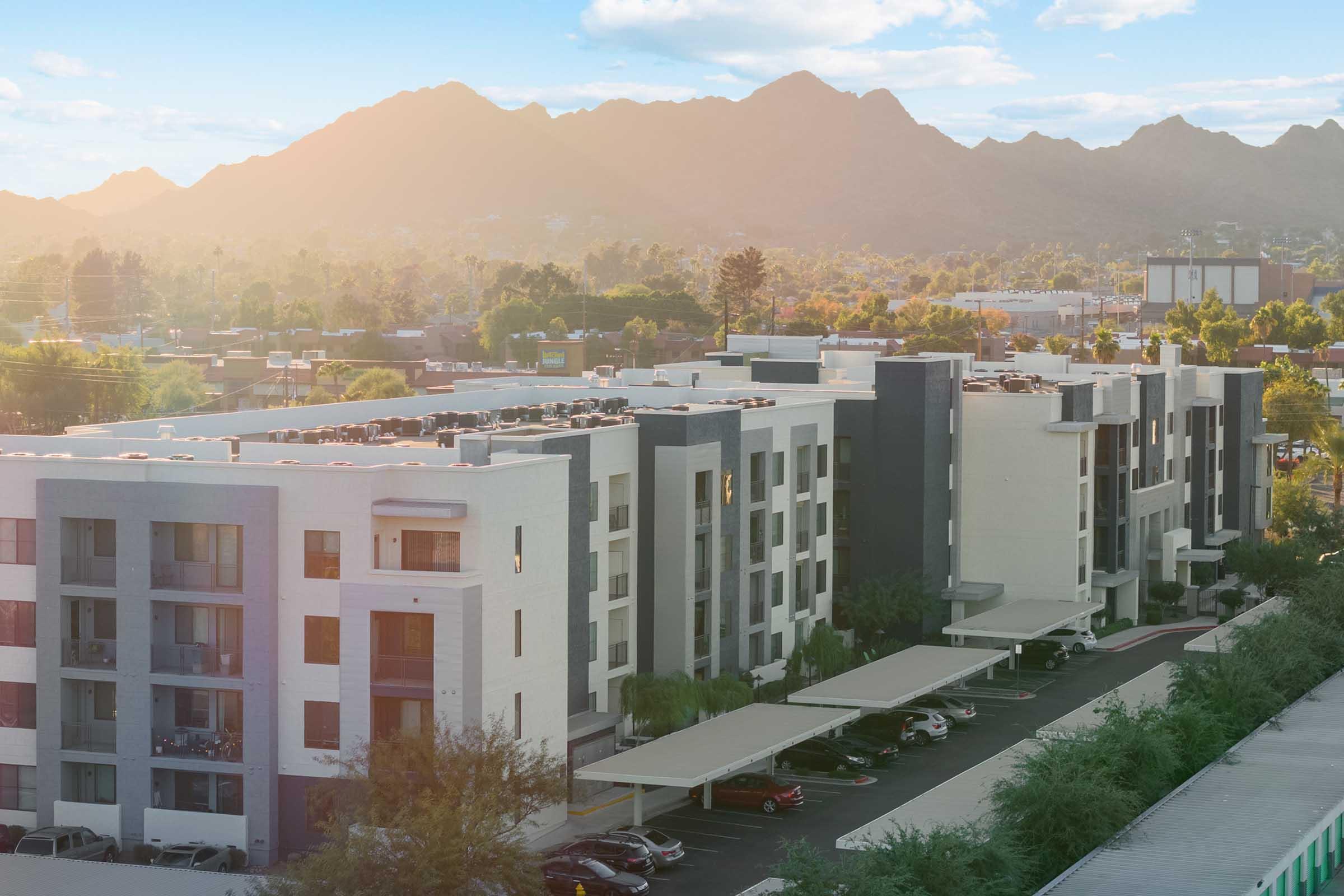 A view of a modern apartment complex with multiple stories, surrounded by trees and parking spaces. In the background, there are mountains bathed in warm sunlight, creating a scenic landscape.