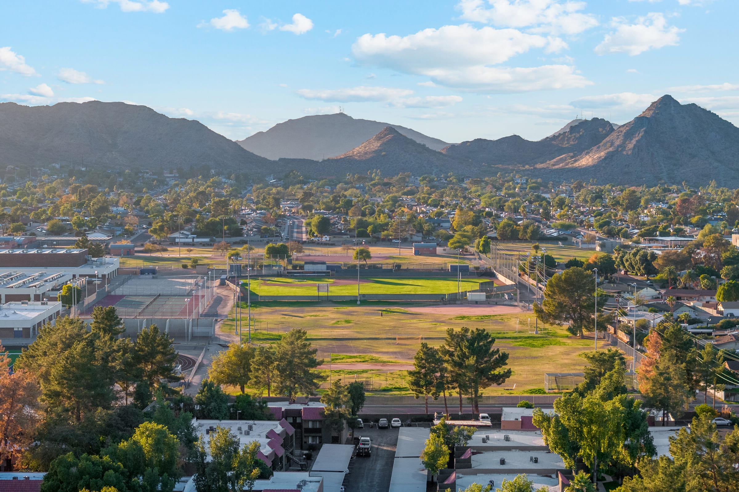 A panoramic view of a sports field surrounded by residential areas and lush greenery, set against a backdrop of mountains under a clear blue sky with scattered clouds. 