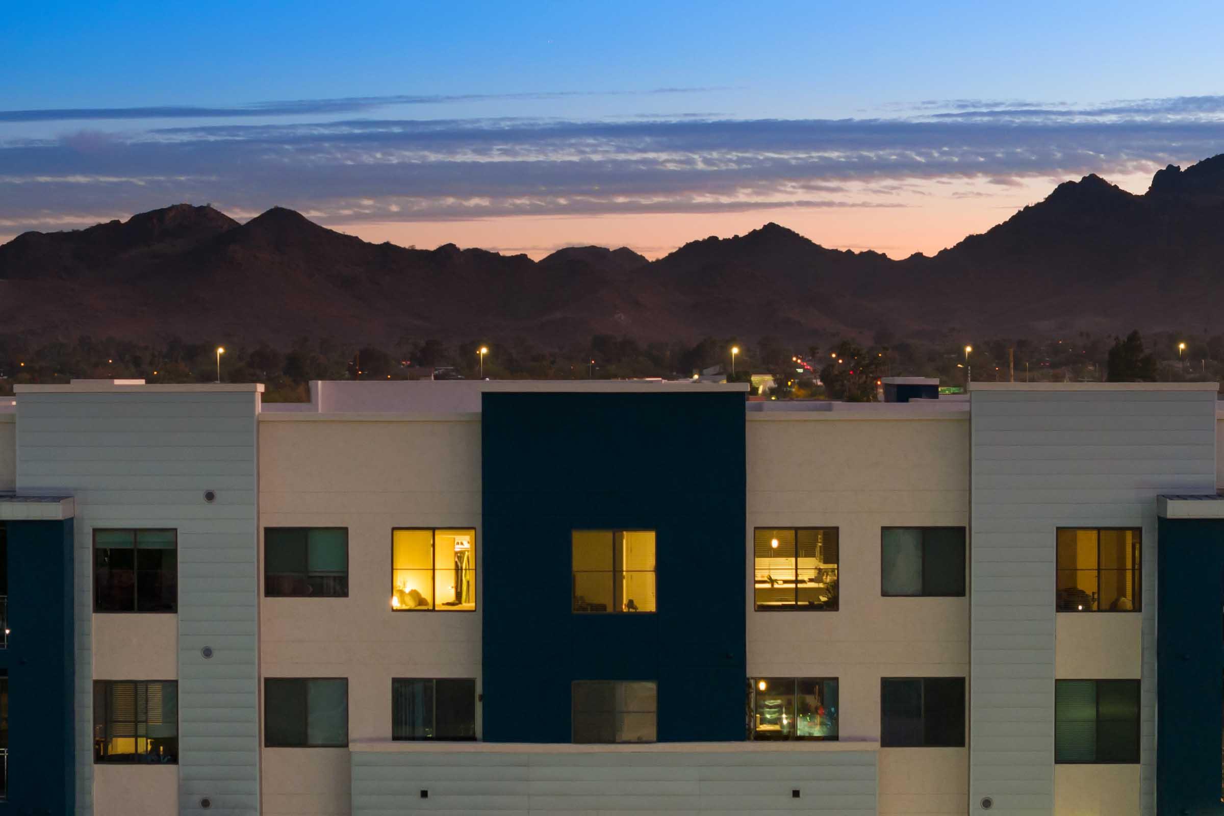 A modern building with large windows is illuminated at dusk, revealing warm light inside. 