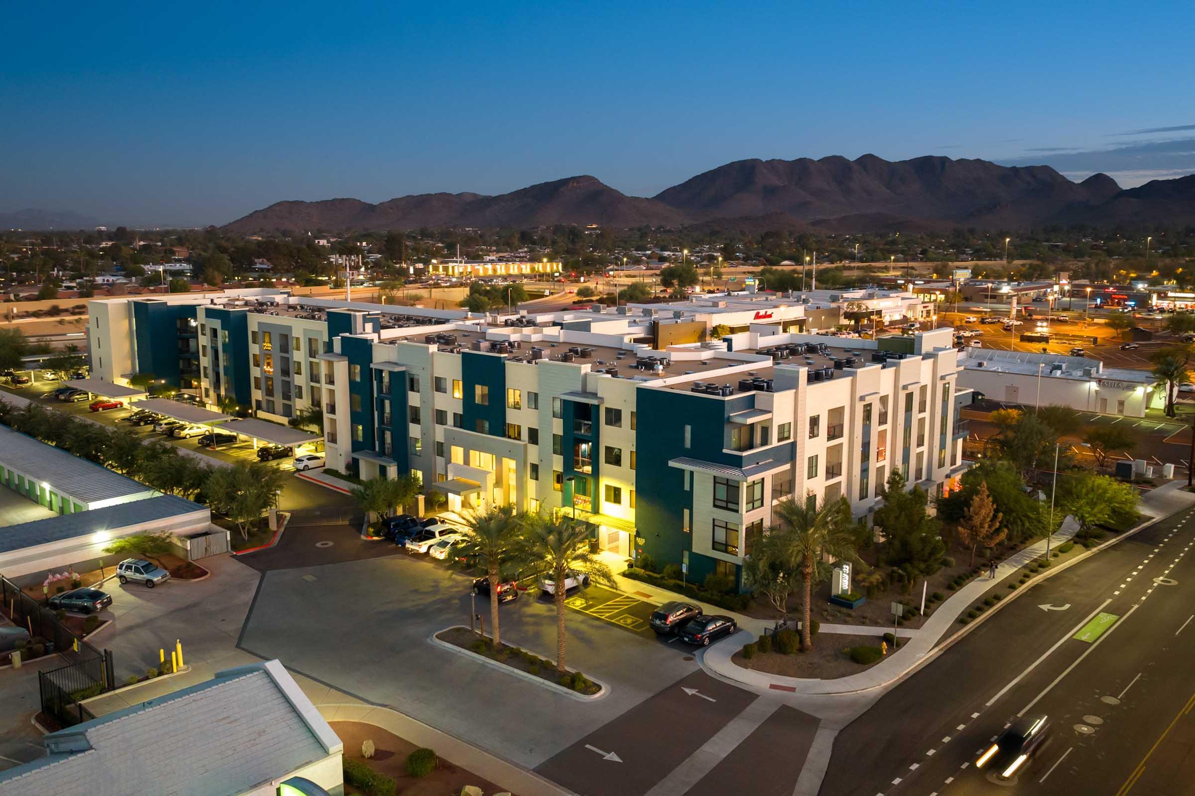 A modern multi-story residential building with a blue and white facade, surrounded by palm trees and parking lots. 