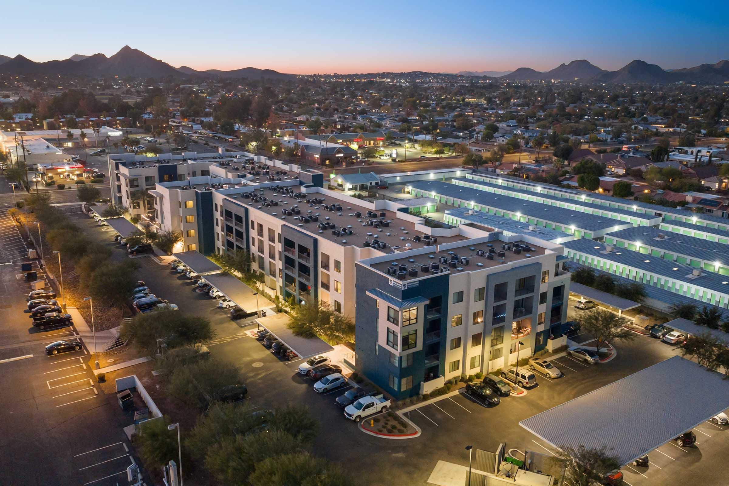 An aerial view of a modern multi-story apartment building at dusk, surrounded by parking lots and palm trees. 