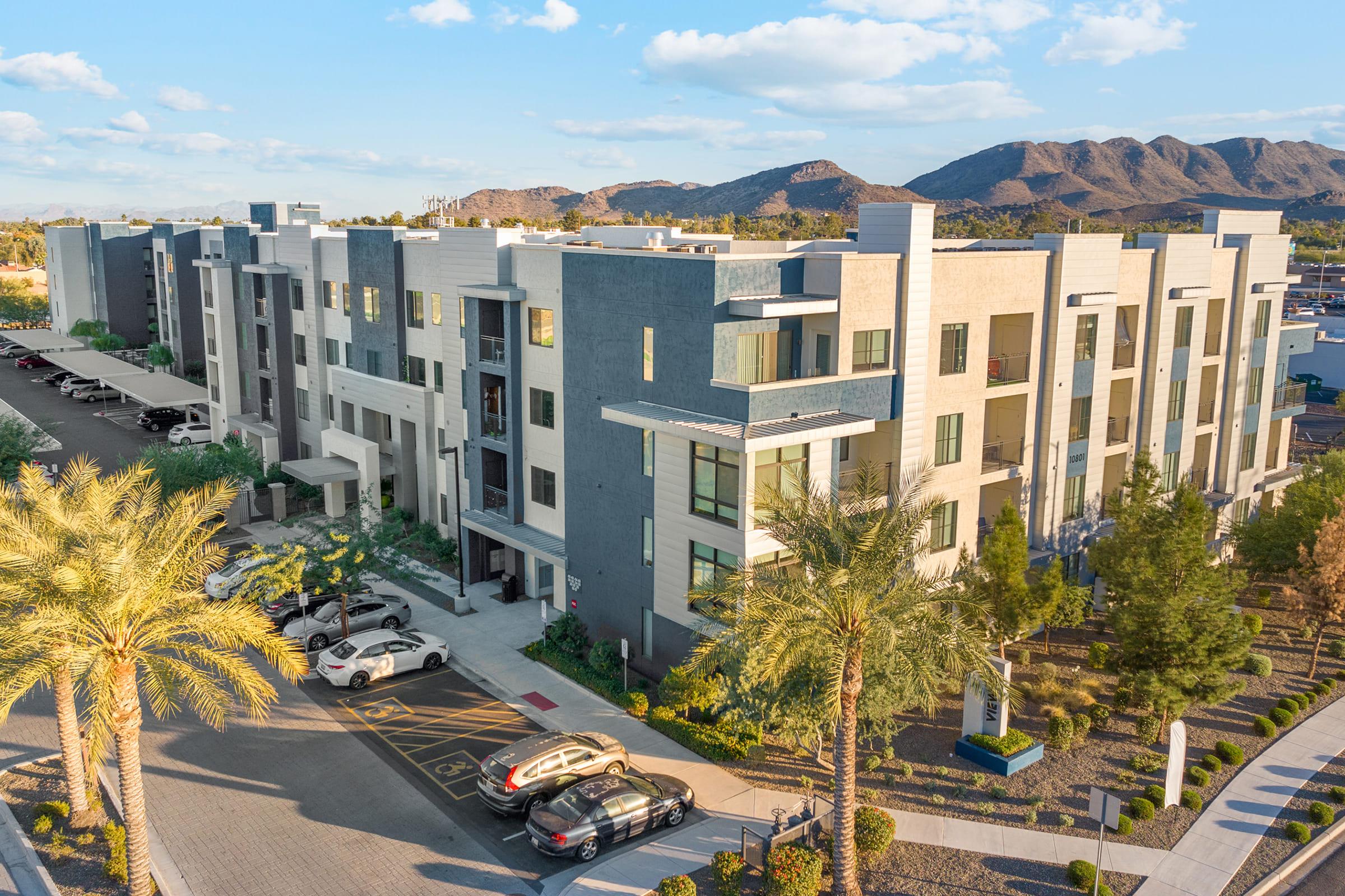 A modern apartment complex surrounded by palm trees and desert landscape, with parking spaces in front. 