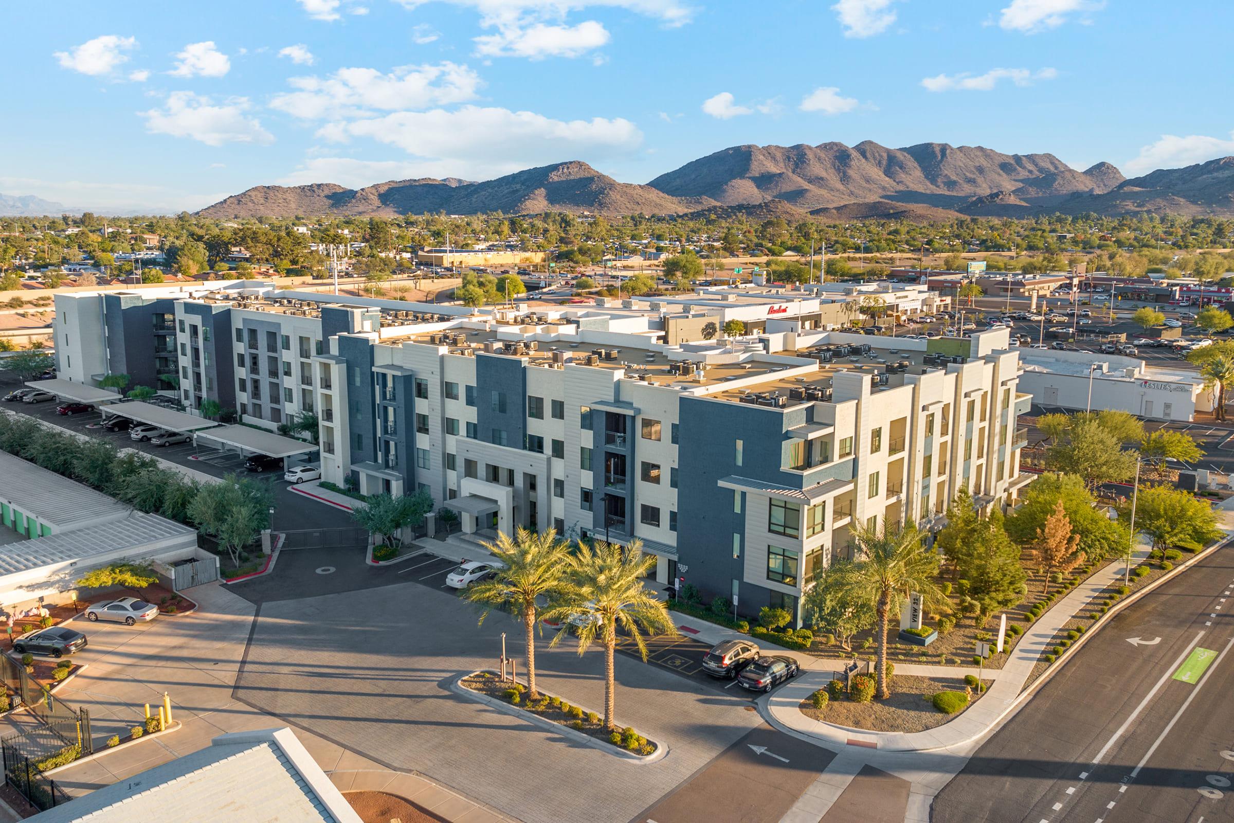 Aerial view of a modern apartment complex surrounded by landscaped greenery and palm trees, with mountains in the background. 