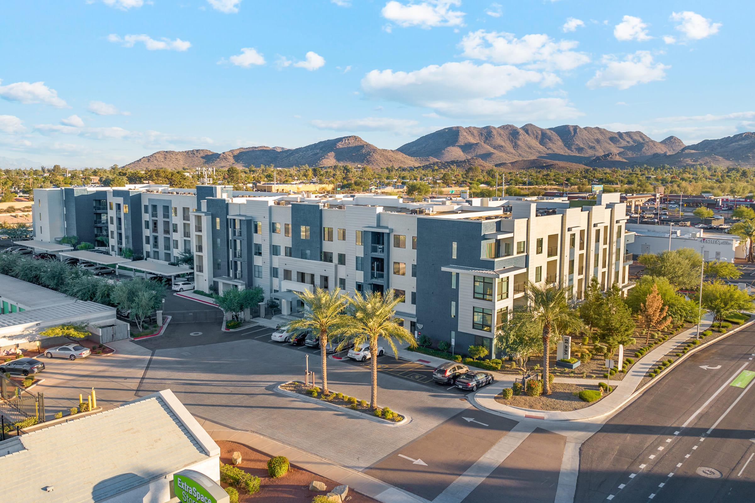 A modern apartment complex surrounded by palm trees and parking spaces, set against a backdrop of mountains and a clear blue sky. 