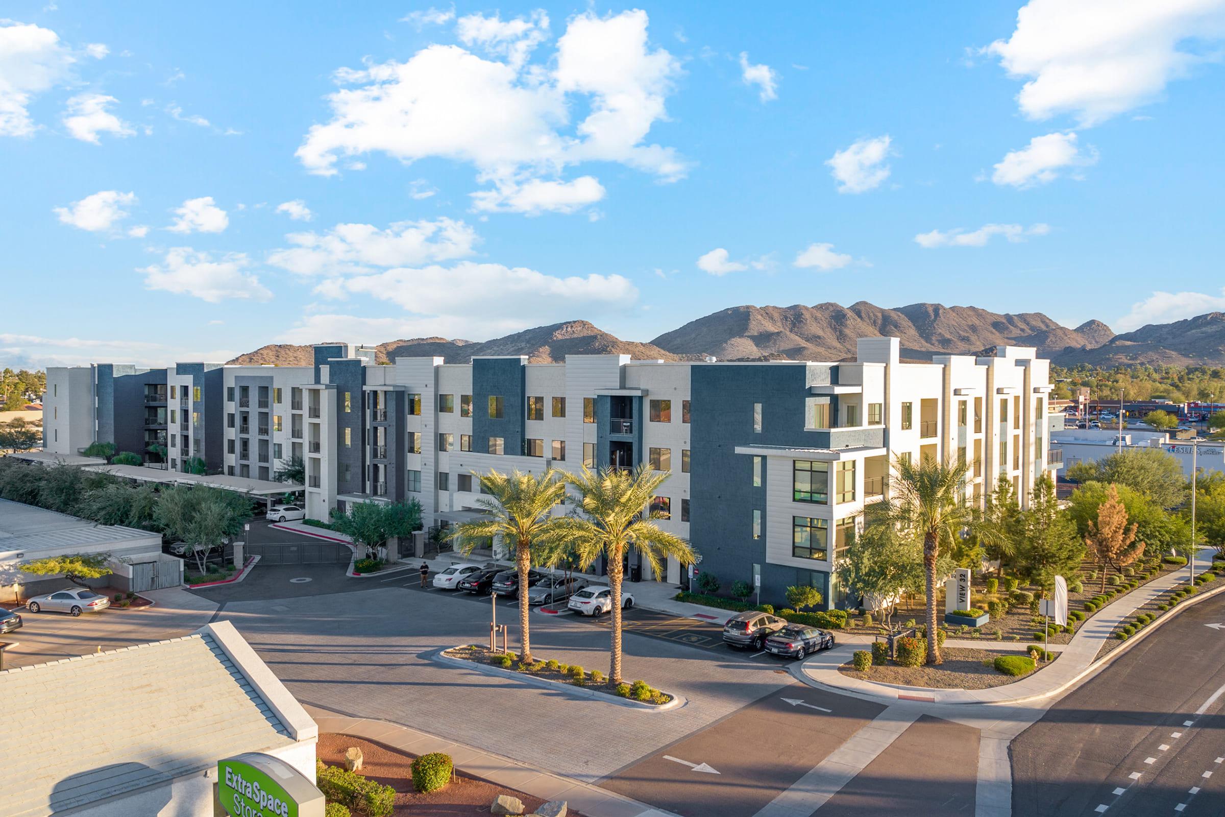 A modern multi-story building with a mix of blue and white exteriors, surrounded by palm trees and a parking area. 