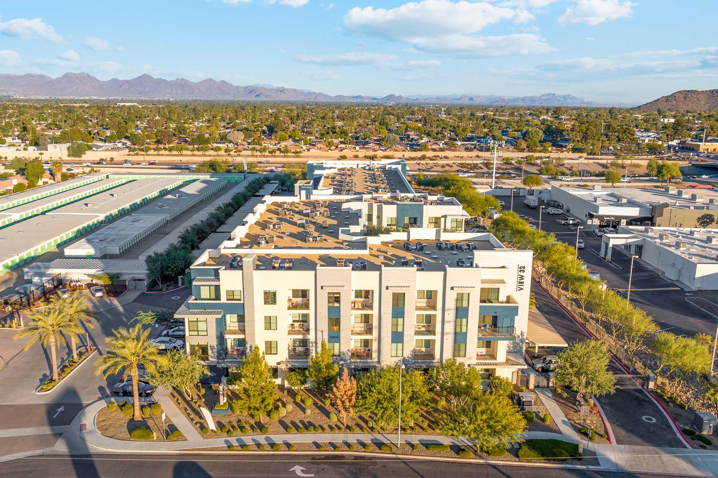 Aerial view of a modern apartment complex surrounded by trees and parking areas, with mountains in the background. 