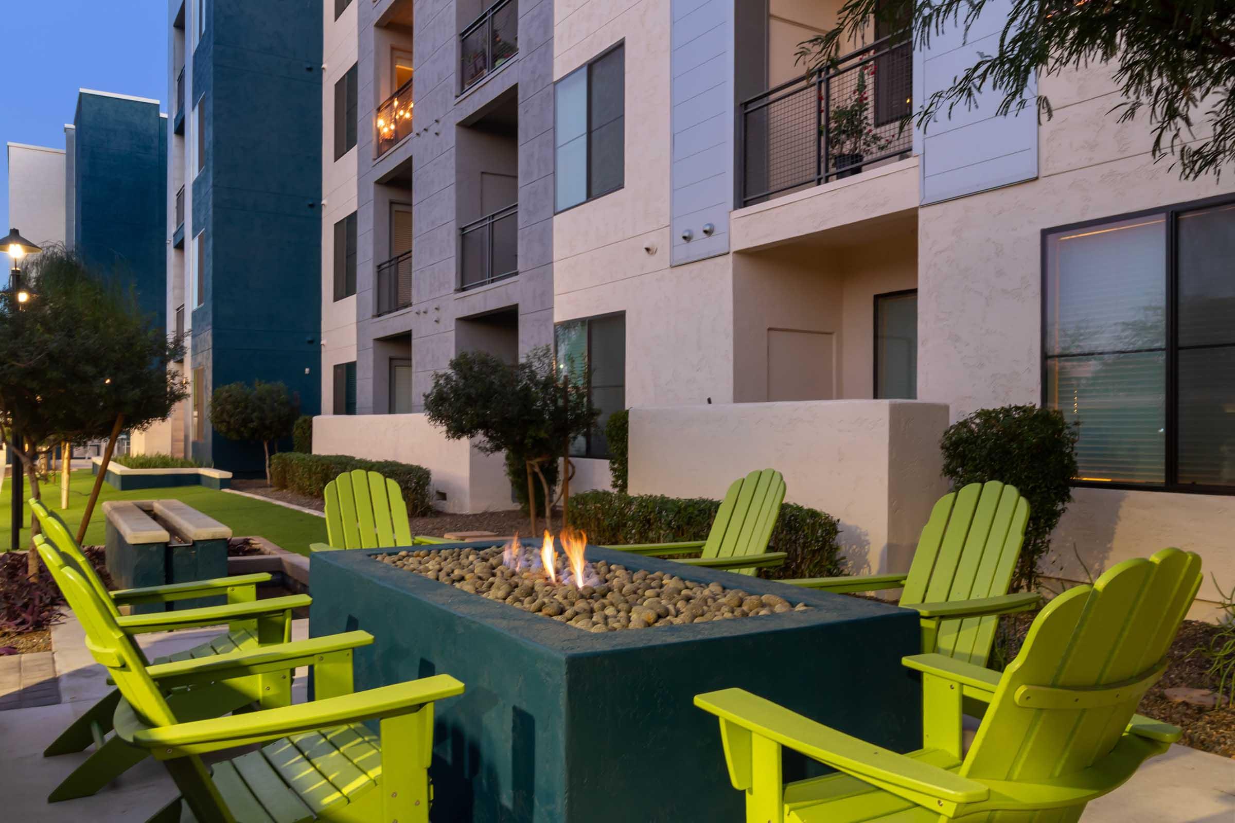 A cozy outdoor seating area featuring bright green Adirondack chairs arranged around a modern fire pit, surrounded by neatly trimmed bushes and an apartment building backdrop in the evening light.