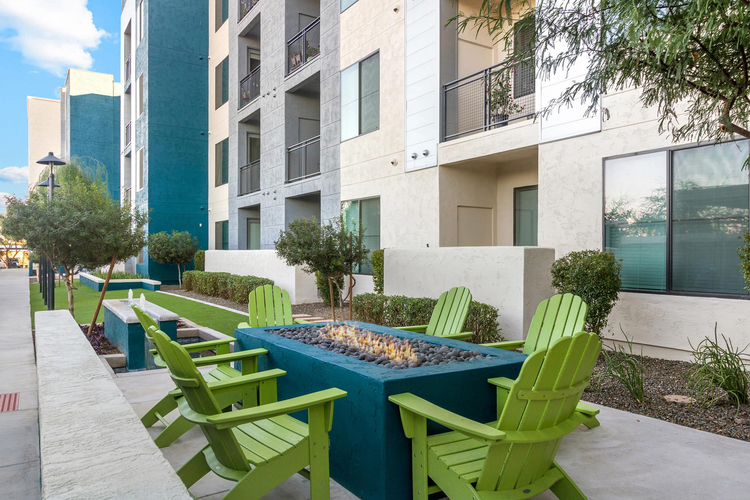 A modern outdoor lounge area featuring a rectangular fire pit with flames, surrounded by green Adirondack chairs. 