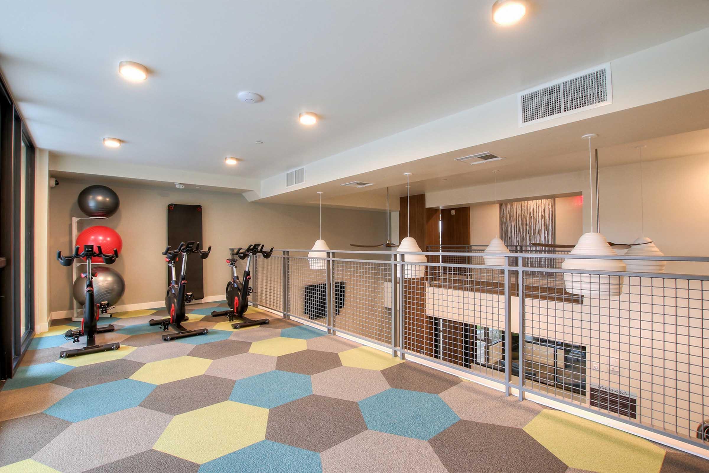 A modern fitness area featuring stationary bikes, exercise balls, and a colorful hexagonal carpet. 