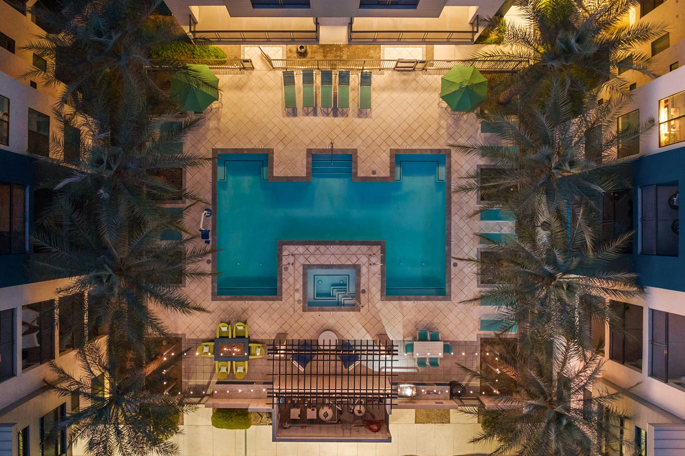An aerial view of a modern outdoor pool area featuring a rectangular pool with surrounding lounge chairs and umbrellas. The space is accented by palm trees and incl