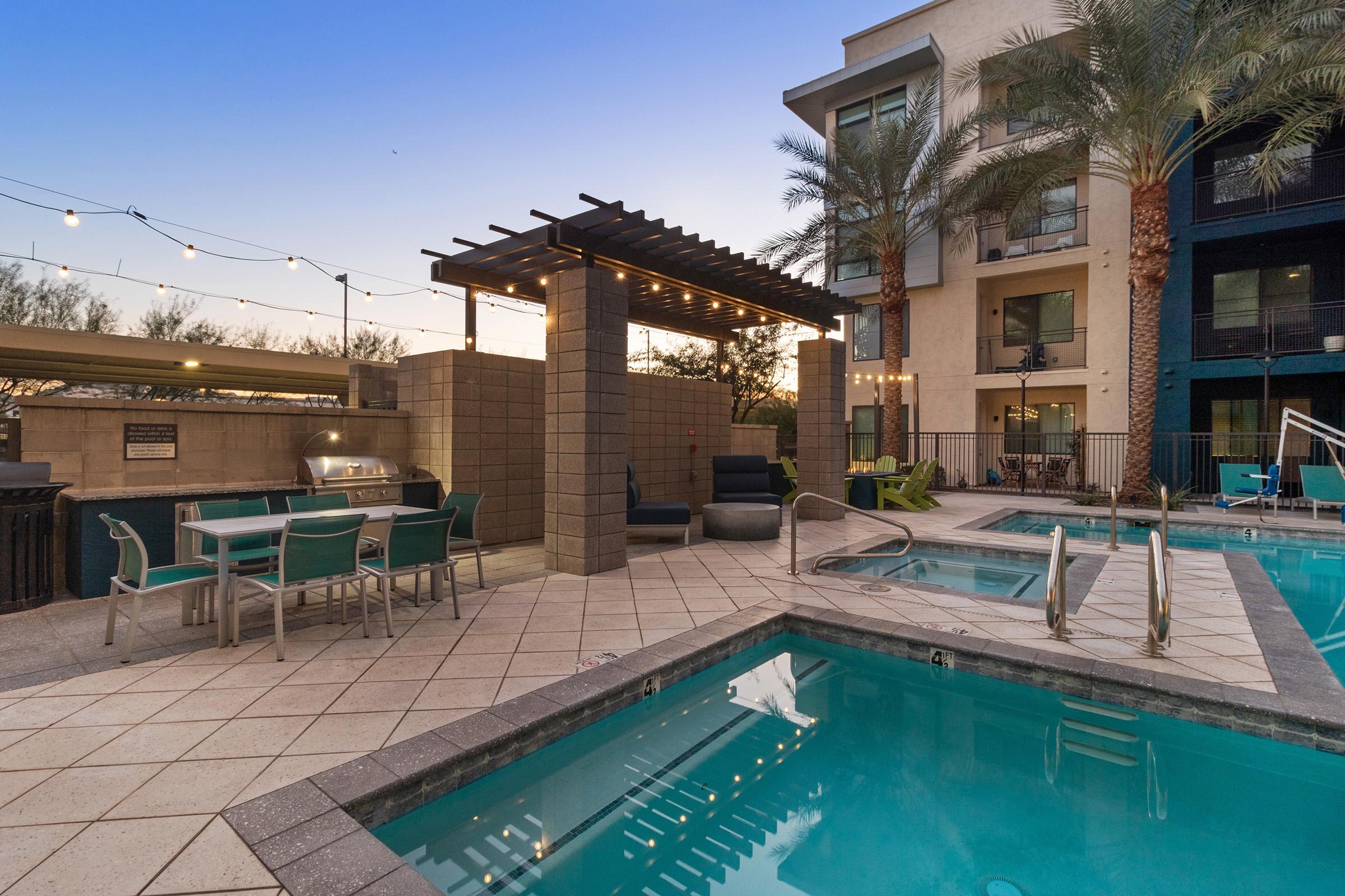 A modern outdoor pool area featuring crystal-clear water, a lounging space with chairs, and a covered grilling station. 